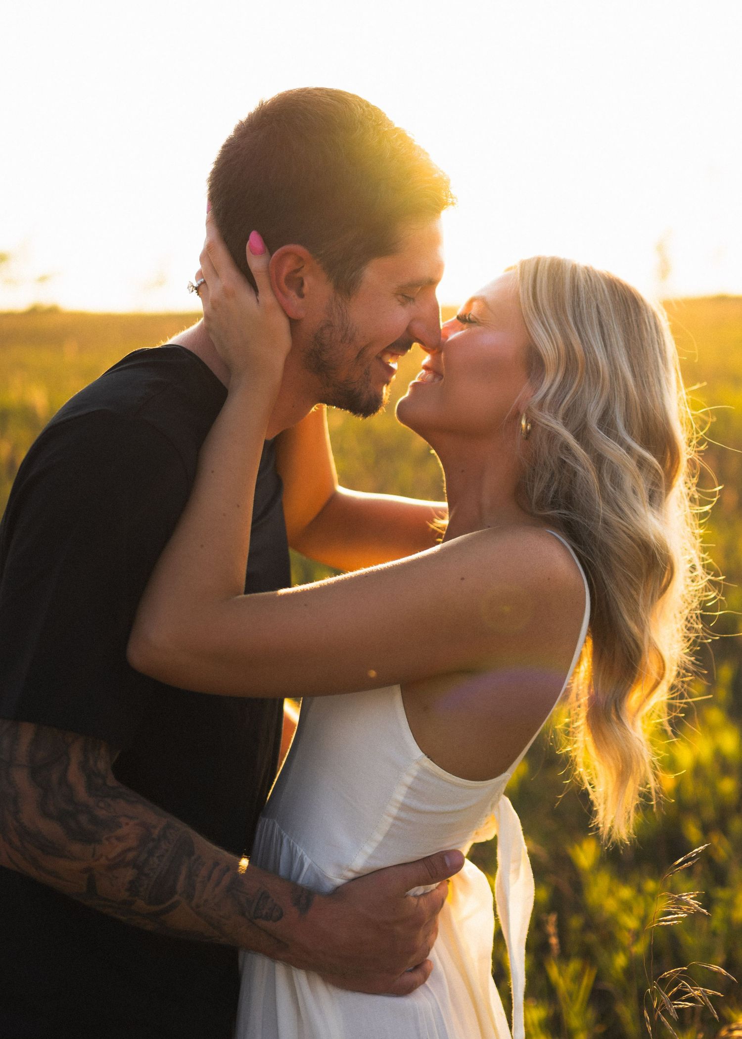 Couple sharing intimate almost-kiss at sunset in field, woman in white dress with tattooed man in black tee.