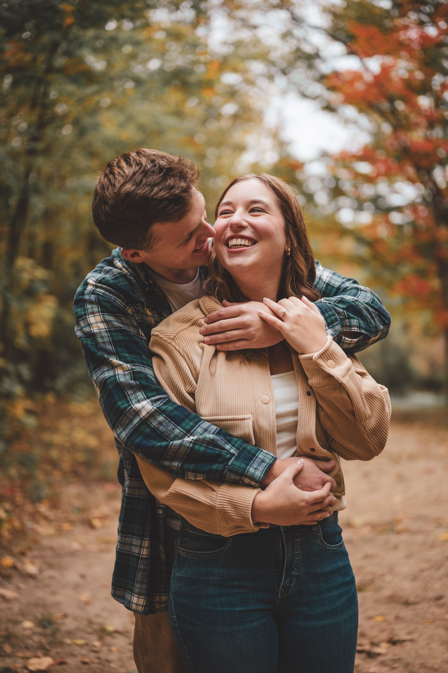 A man embraces a laughing woman from behind on an autumn forest path surrounded by colorful fall foliage.