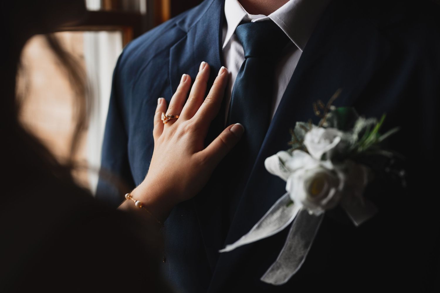 Close-up of bride placing hand on grooms navy suit jacket with white rose boutonniere.