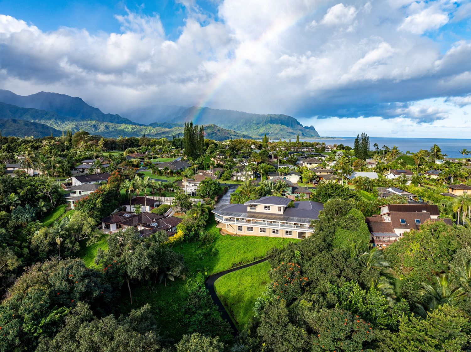 Aerial view of a lush tropical neighborhood with ocean views, mountains, and a rainbow in Kauai, Hawaii.