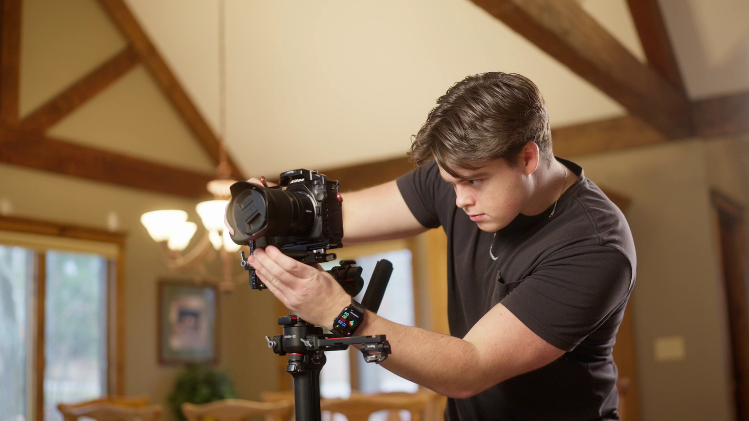 Young man in black shirt adjusting a DSLR camera on a tripod indoors with vaulted wooden ceiling.
