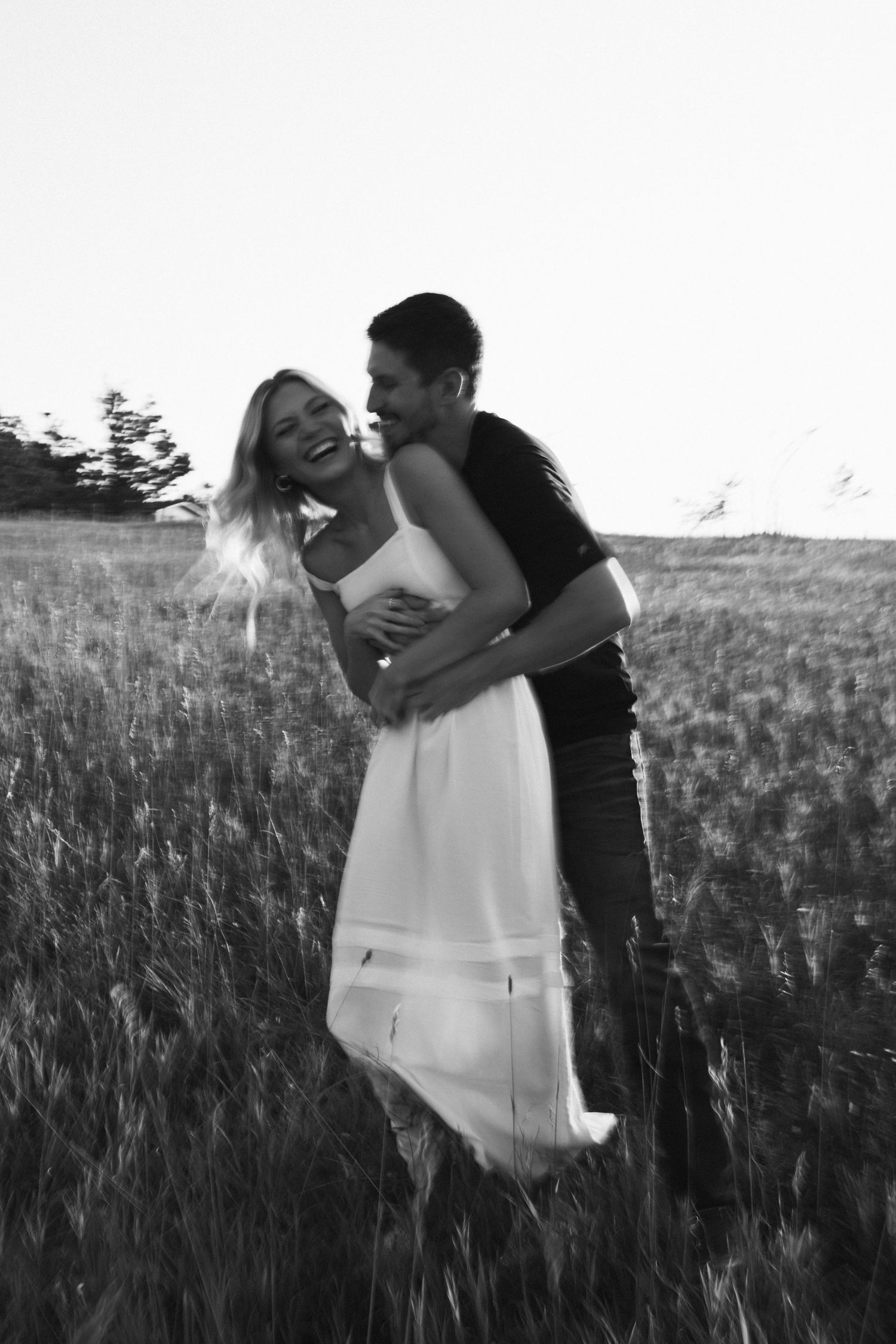 Black and white photo of joyful couple embracing in field, woman in white dress laughing as man holds her close.