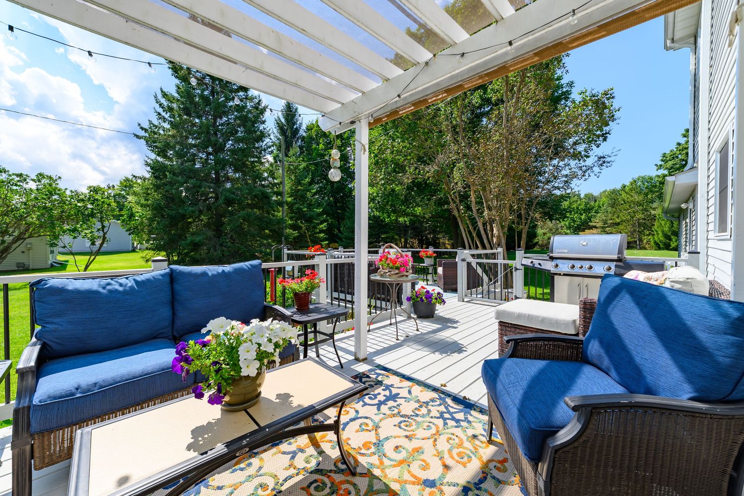Outdoor deck with blue cushioned furniture, pergola, string lights, and lush garden views.