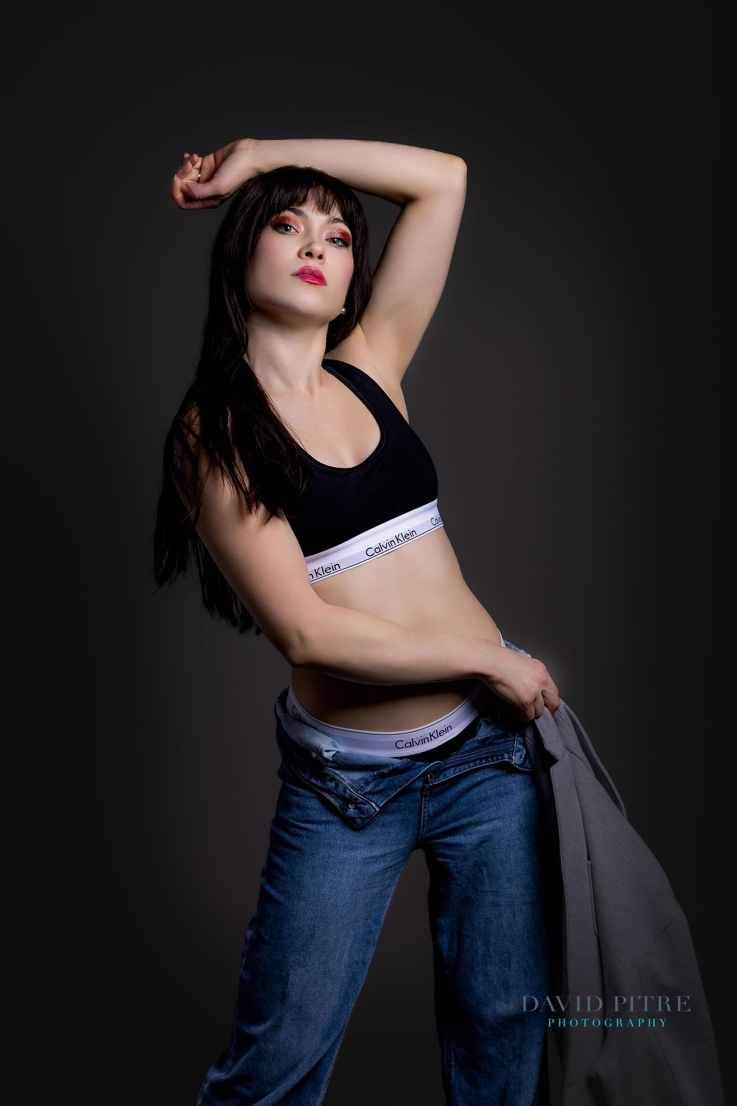 Fashion model in black top and jeans poses against dark background in artistic studio photoshoot series.