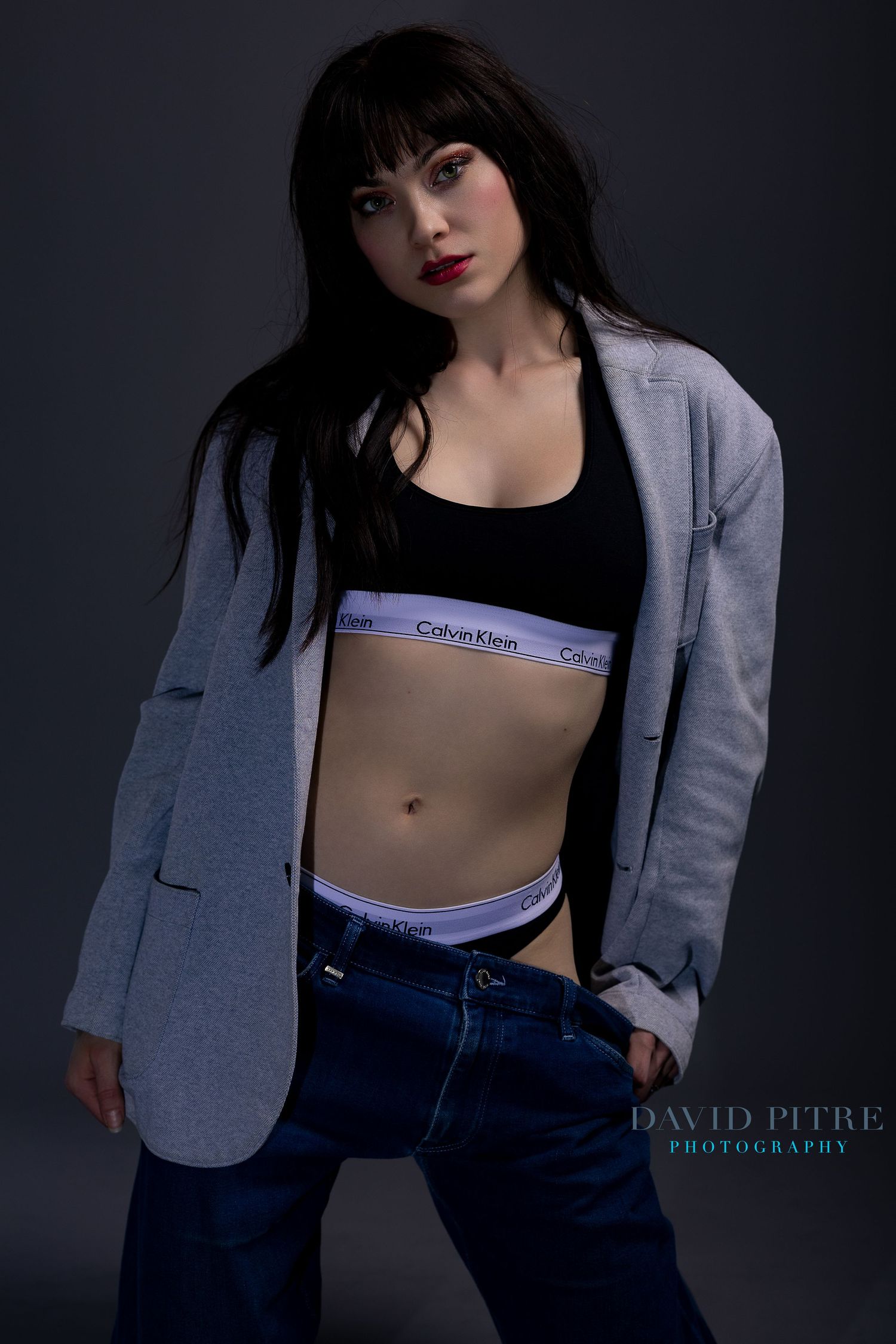 Fashion portrait series with model wearing grey blazer, Calvin Klein top and denim in dark studio.