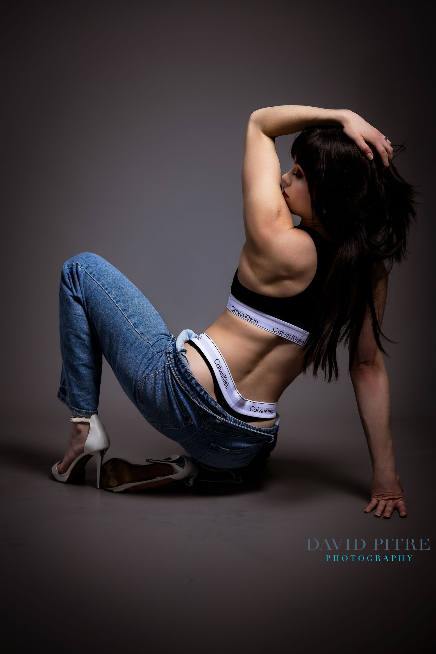 Fashion model in Calvin Klein underwear and jeans poses dramatically against dark background in studio setting.