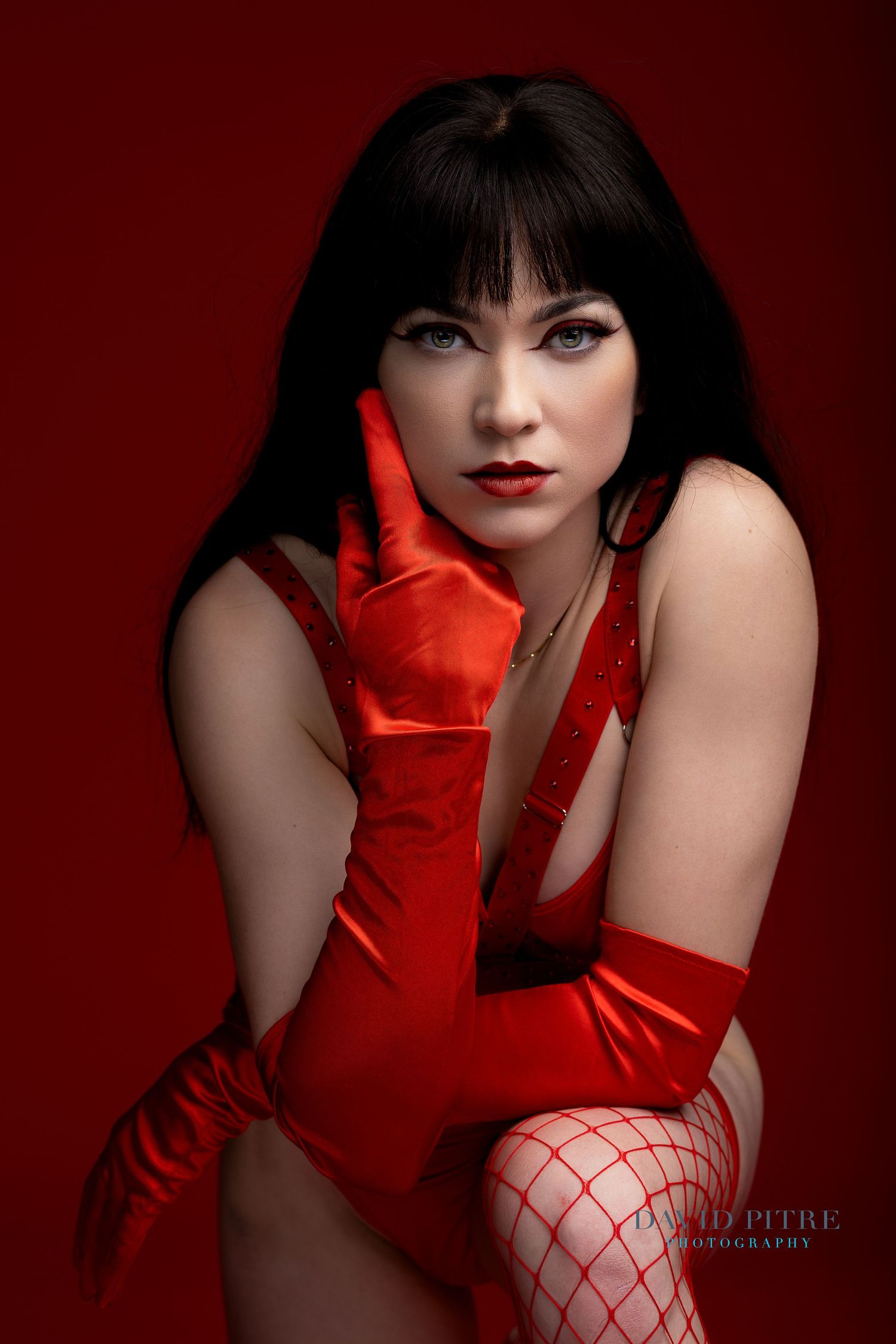 Glamorous portrait against red background featuring bright red satin gloves, bodysuit and fishnet stockings in dramatic lighting.