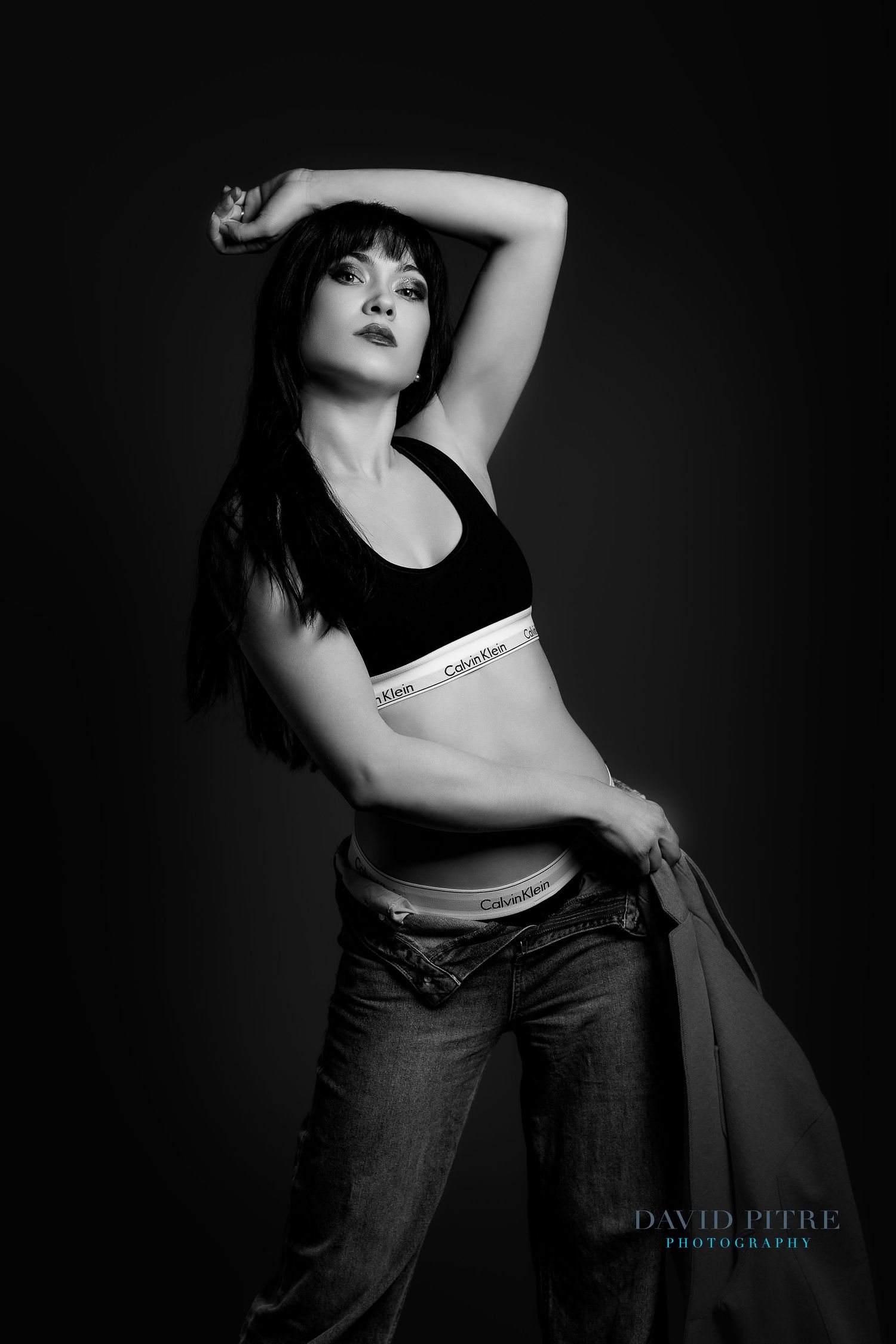 Black and white fashion portrait of a woman in Calvin Klein sports bra and jeans posing against dark background.