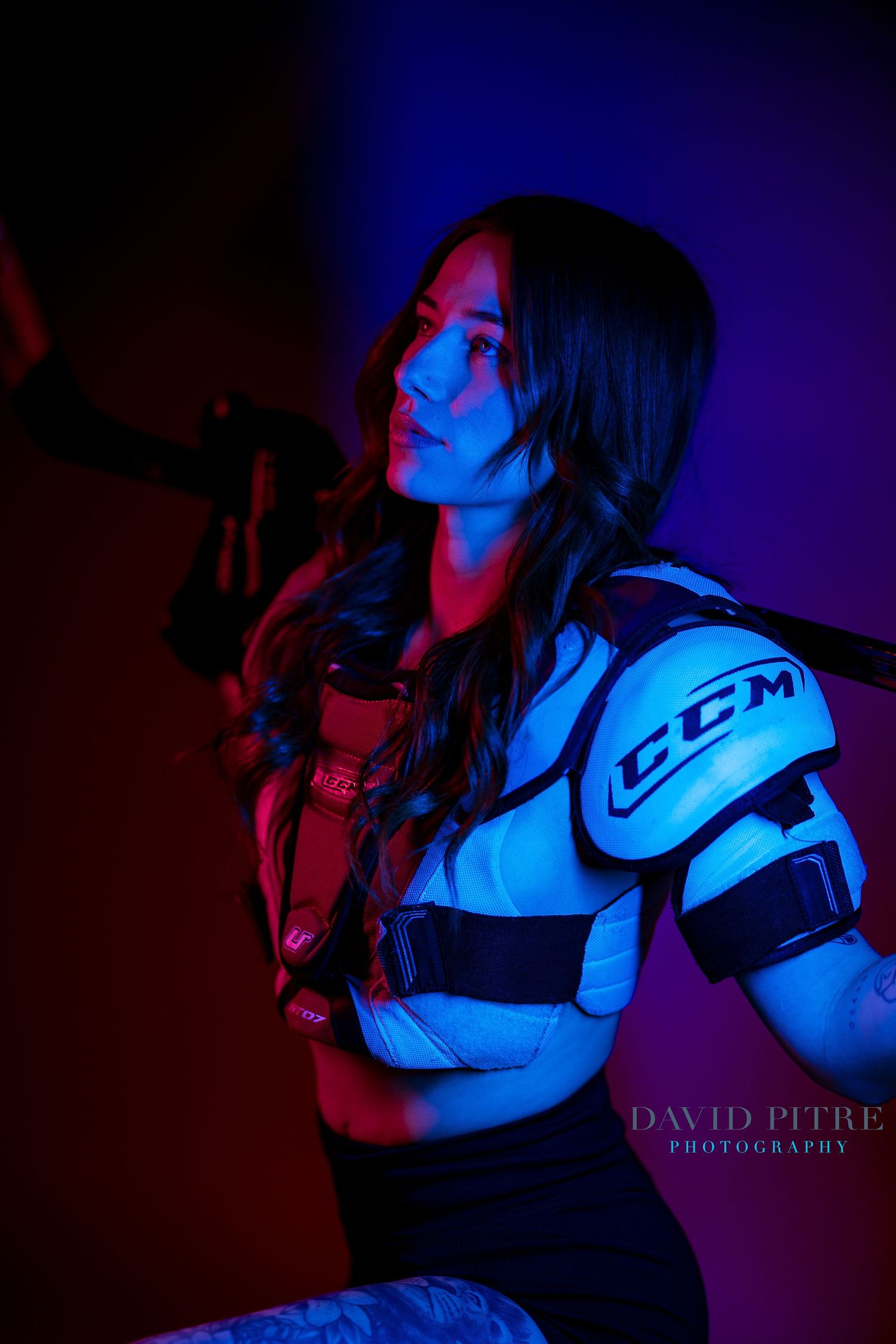 Dramatic portrait in neon lighting shows person wearing CCM hockey shoulder pads against red and blue background.