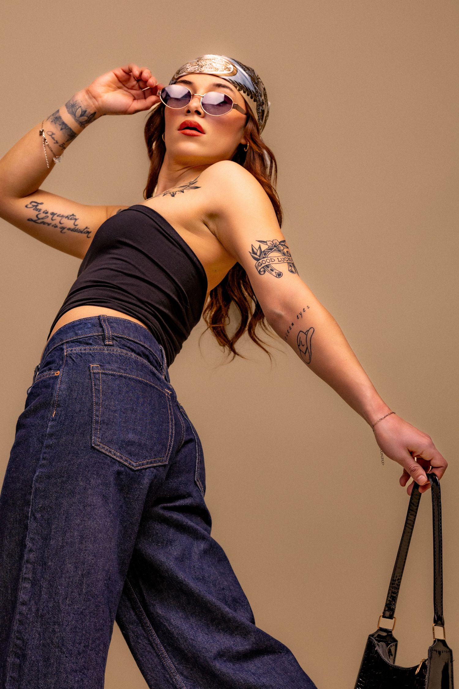 Fashion model in black top and dark jeans poses dynamically against beige background with handbag and sunglasses.