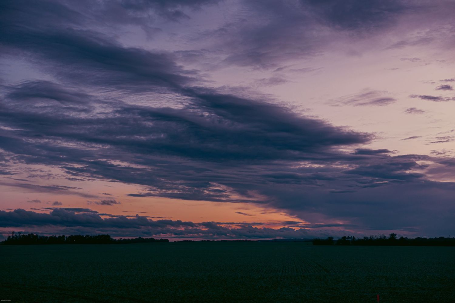 Dramatic panoramic view of dark storm clouds at sunset over a rural landscape with silhouetted trees on the horizon.