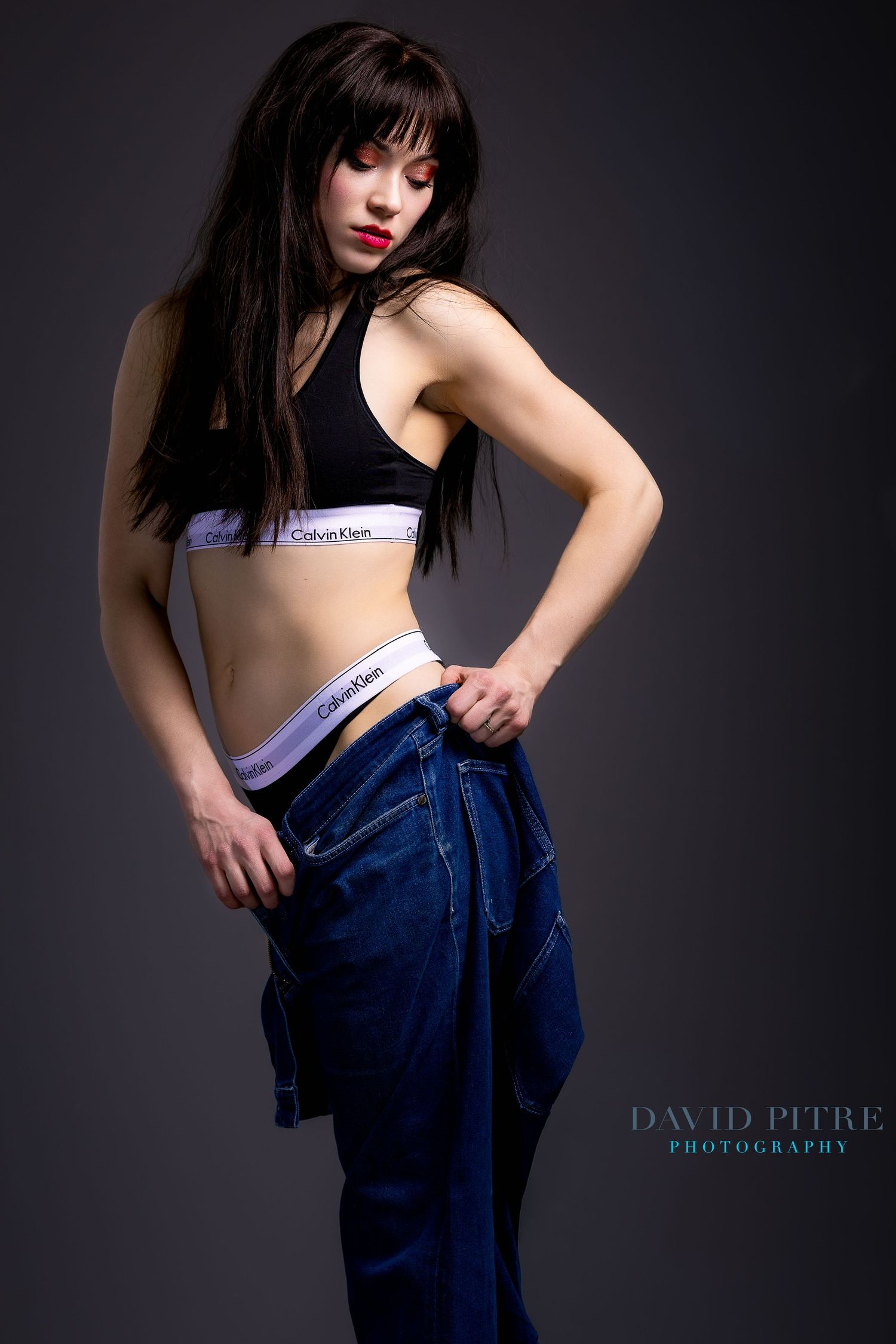 Fashion model wearing Calvin Klein sports bra and denim jeans poses against dark background in artistic photoshoot series.