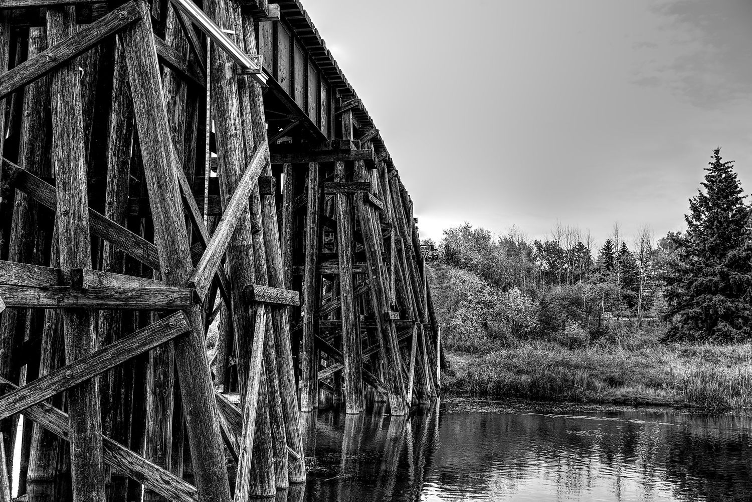 Black and white photograph of an old wooden railroad trestle bridge reflected in calm water below.