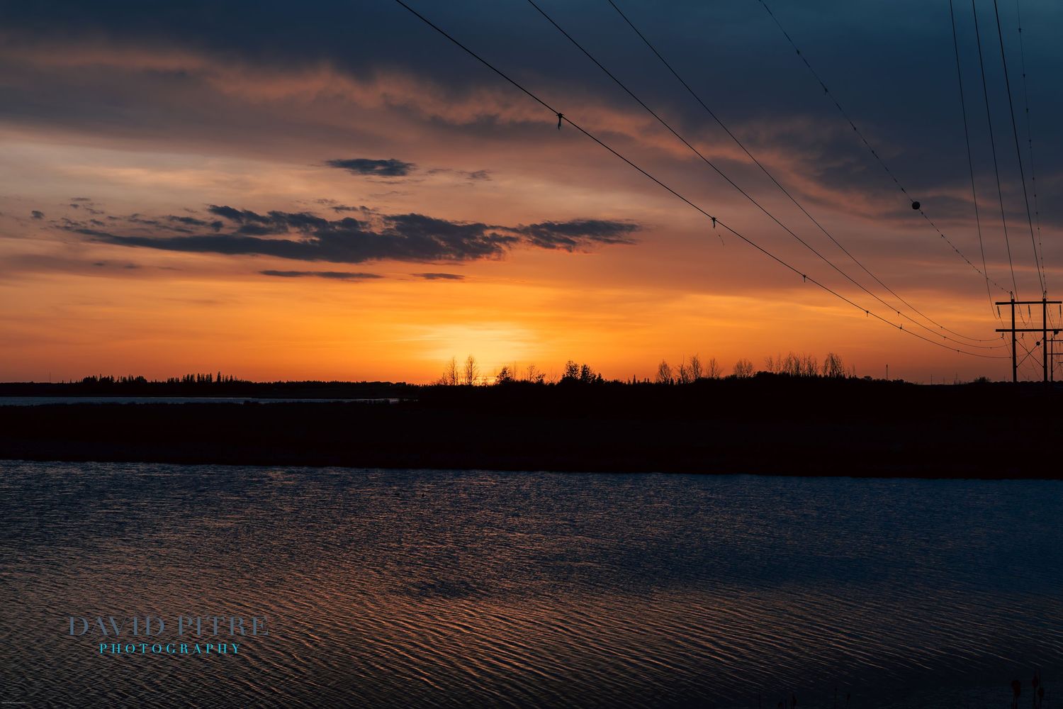 Dramatic sunset silhouettes power lines and trees against a vibrant orange and purple sky over a rural landscape.