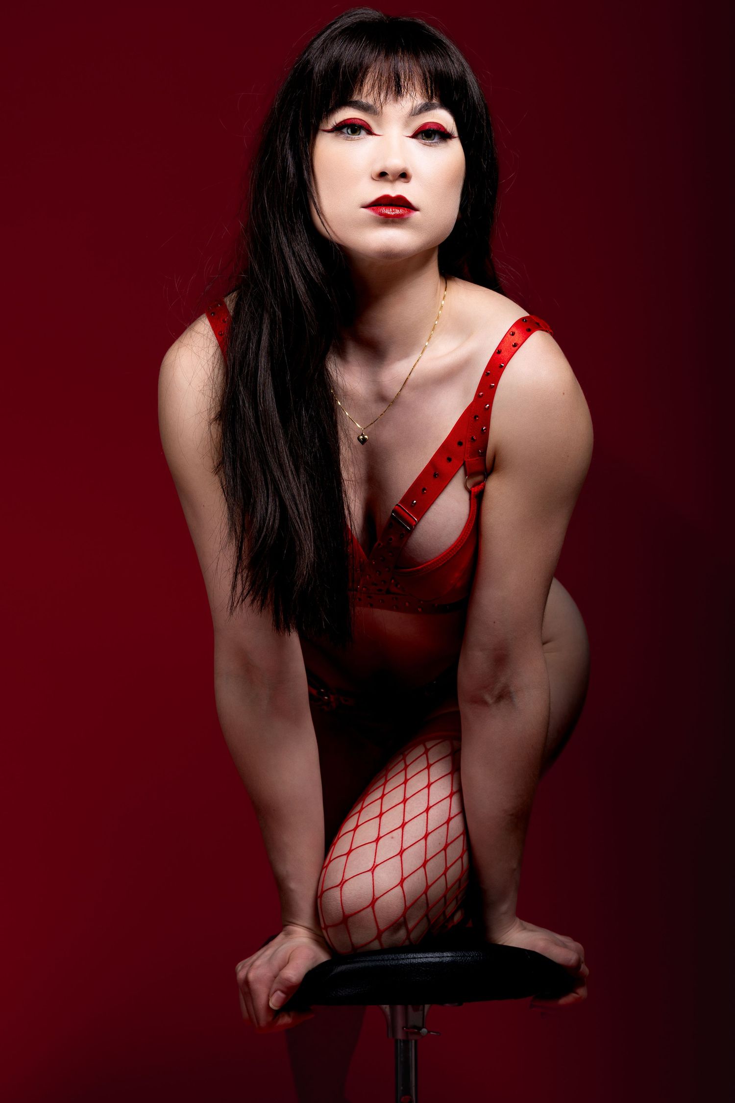 A model in a red dress and fishnet stockings poses on a stool against a deep burgundy background.