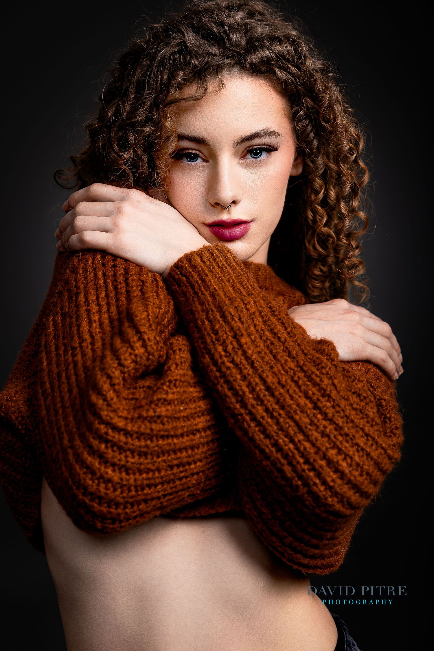 Artistic portrait featuring curly hair and a chunky brown knit sweater against a dark background.