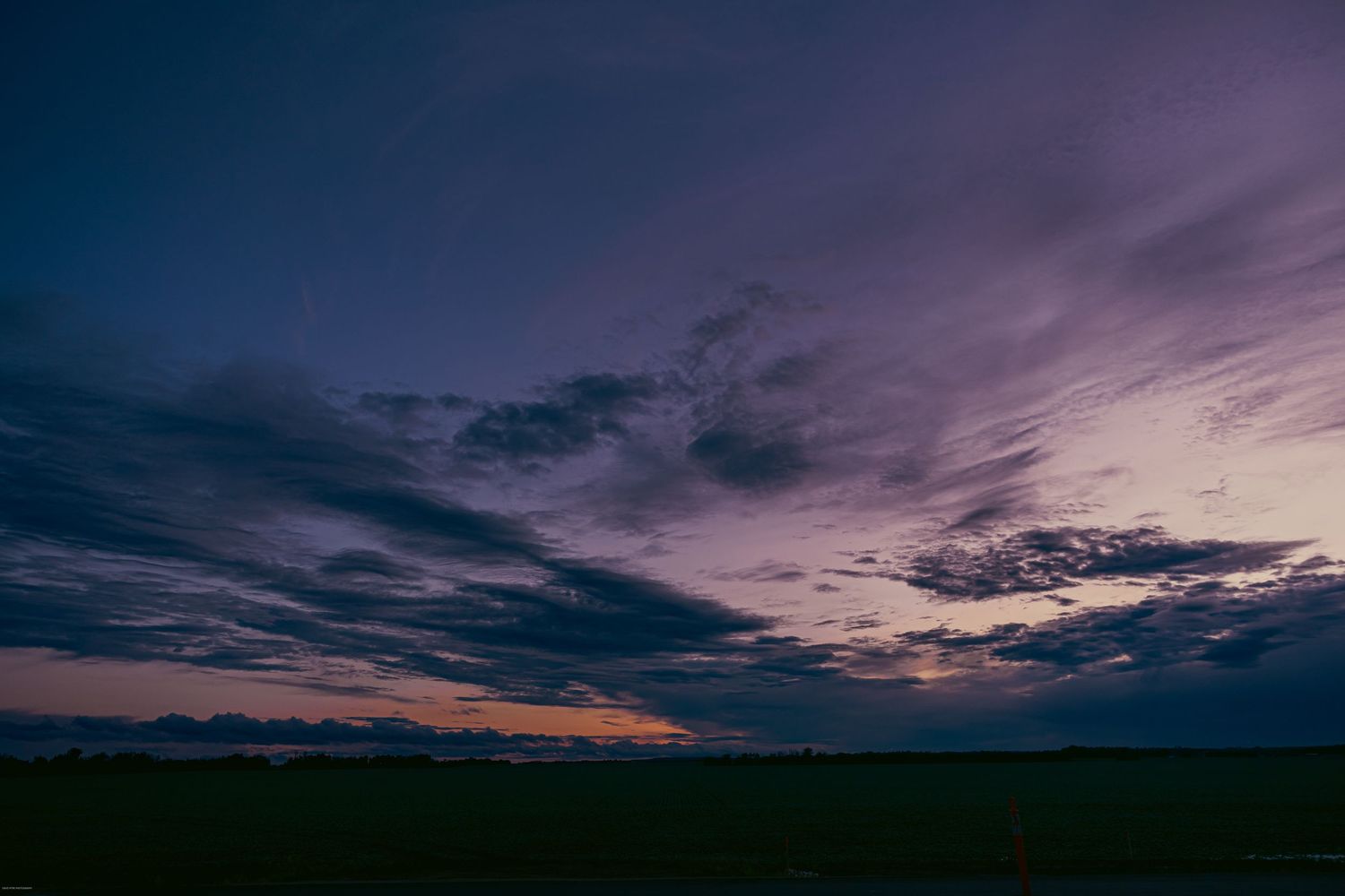 Dramatic purple and blue sunset sky over a dark silhouetted rural landscape with scattered clouds at dusk.