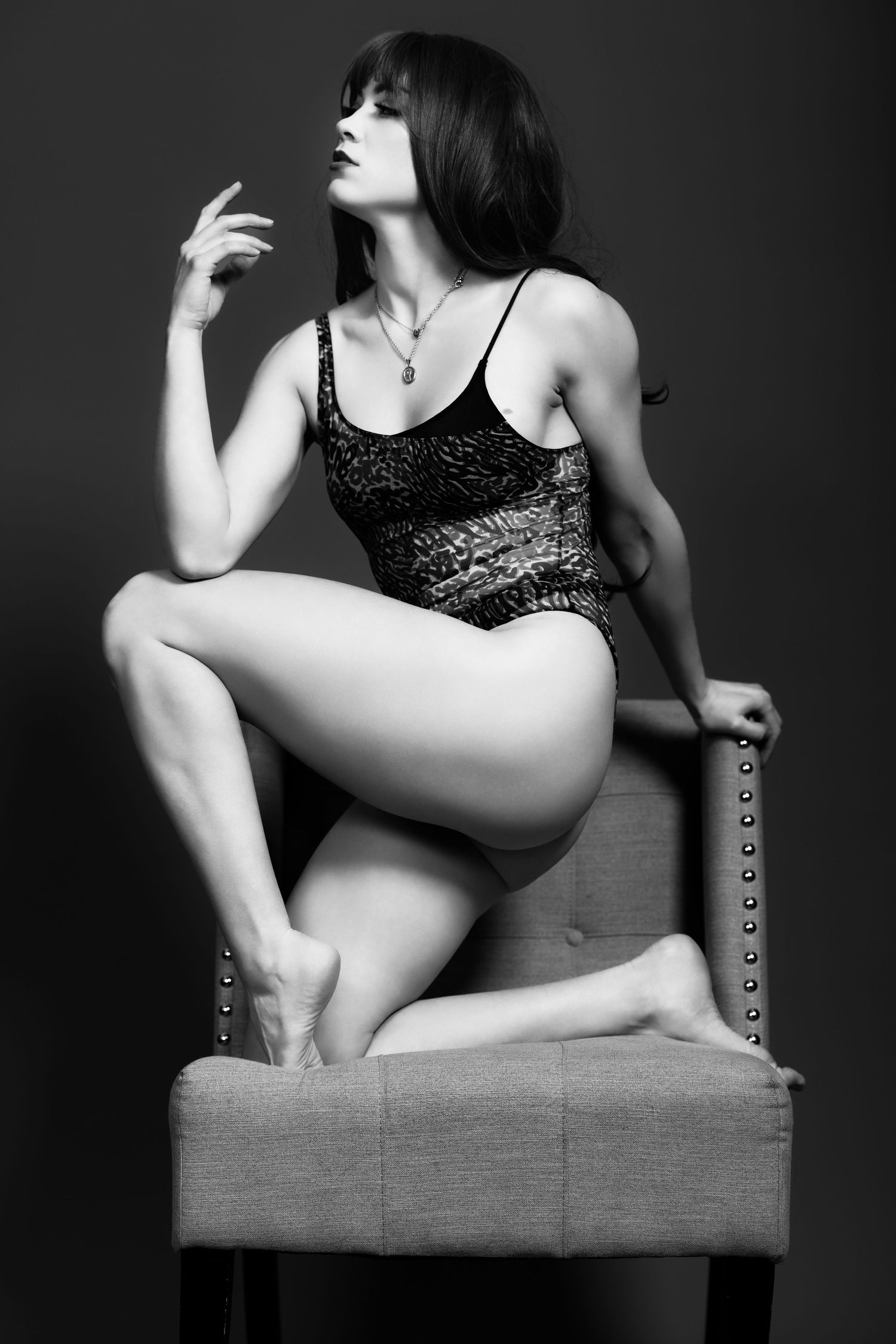Artistic black and white boudoir photo of a figure in lace bodysuit posing dramatically on an upholstered chair.