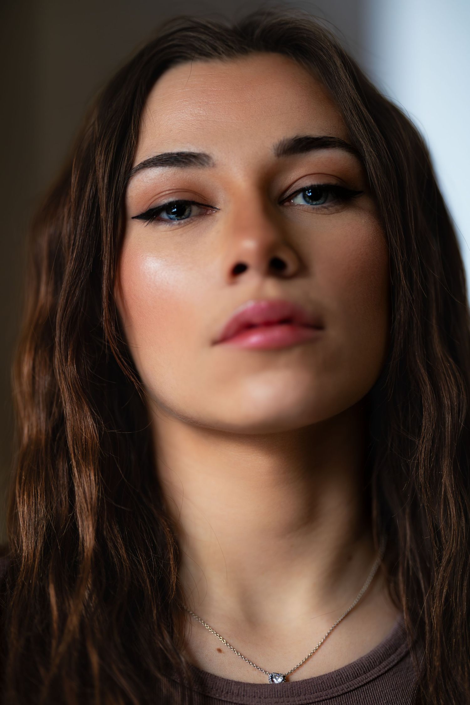 Close-up portrait of someone with long brown hair, pink lipstick and smoky eye makeup looking upward against dark background.