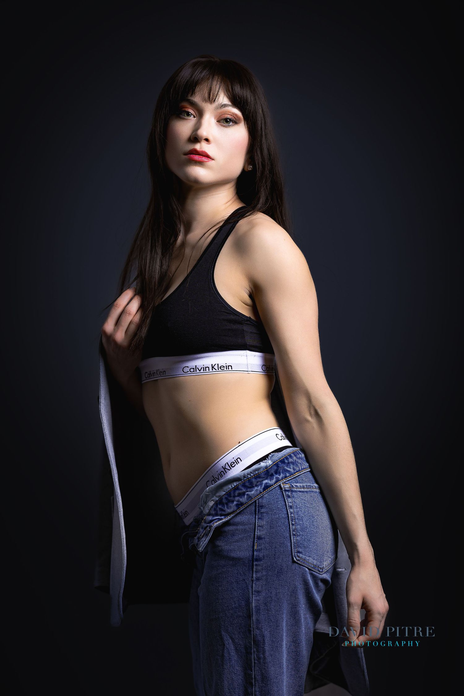 Fashion model in Calvin Klein sports bra and jeans poses against dark background in moody studio lighting.