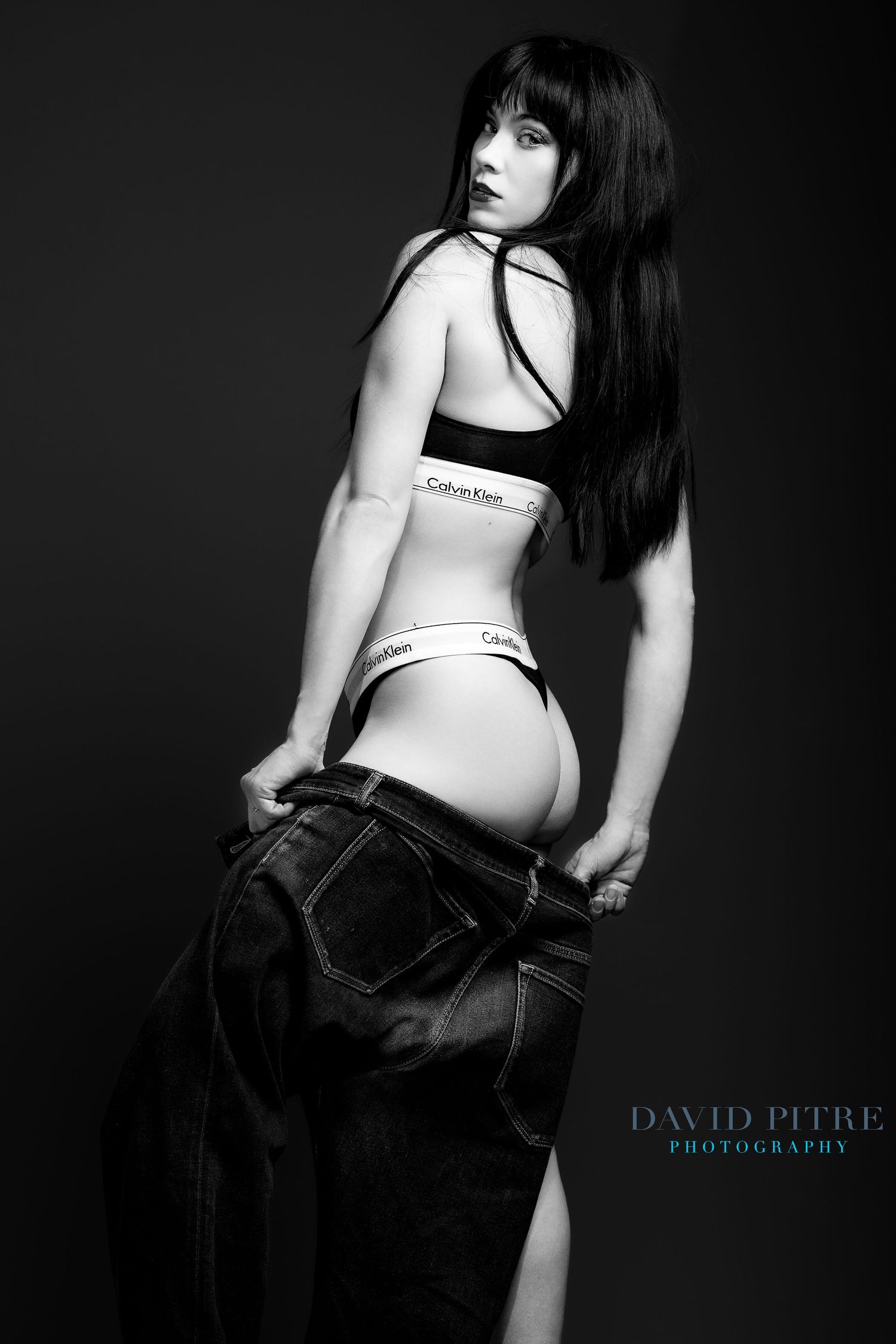 A black and white artistic maternity photoshoot showing a pregnant figure in Calvin Klein underwear against dark backdrop.