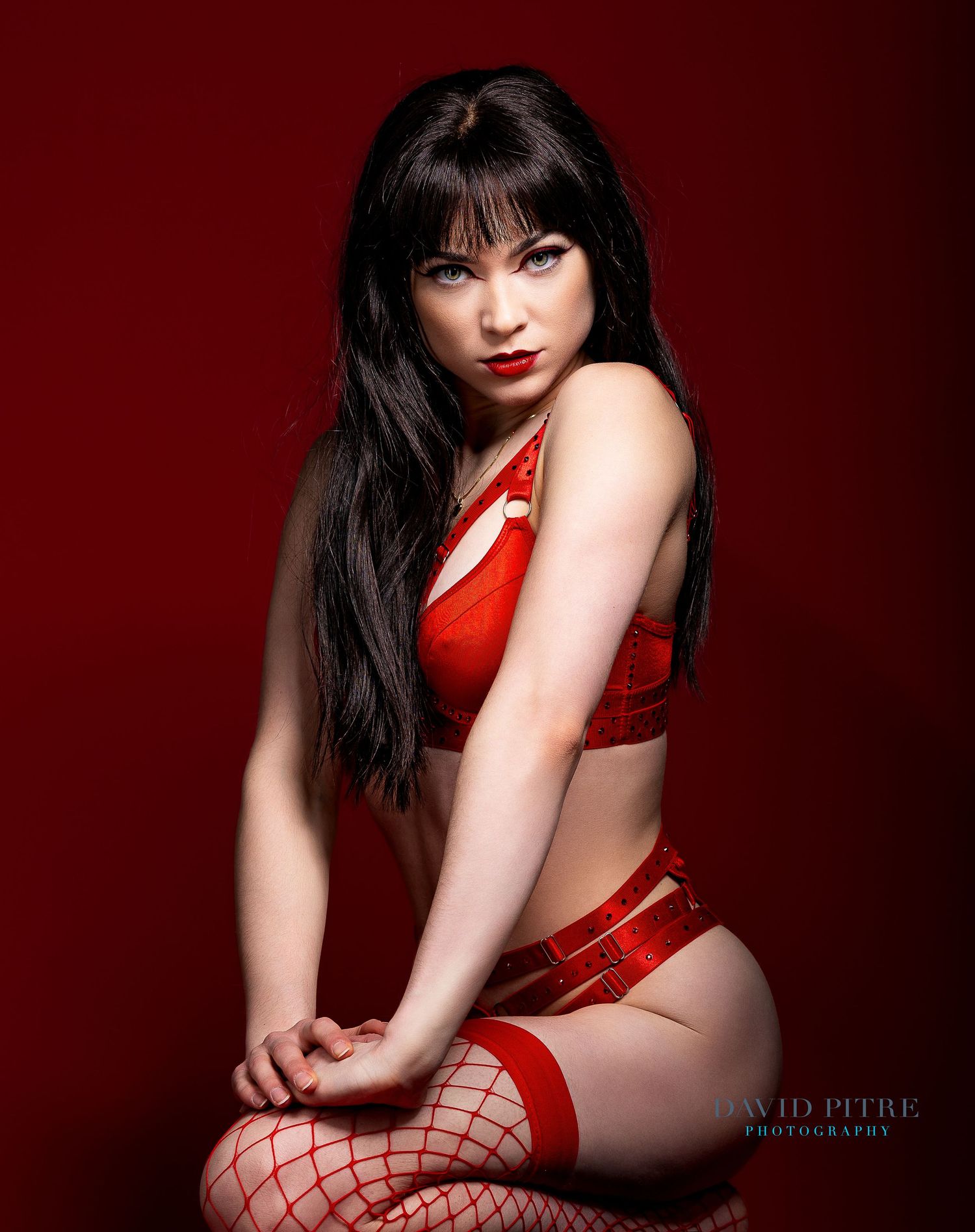 Woman in red lingerie and fishnet stockings poses against dark red background in dramatic boudoir photo.