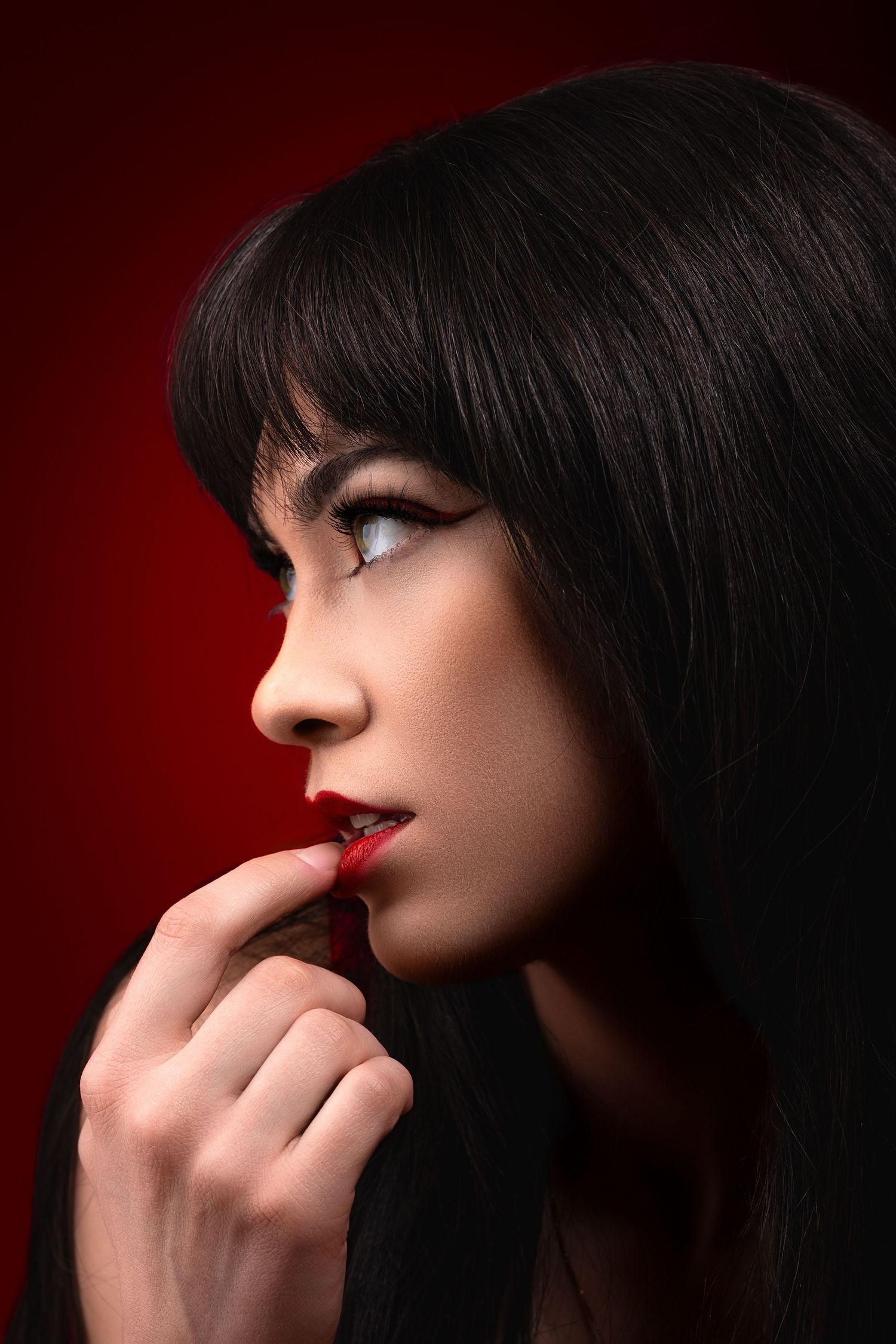Side profile portrait of a woman with dark bangs and red lipstick against a dramatic red background.
