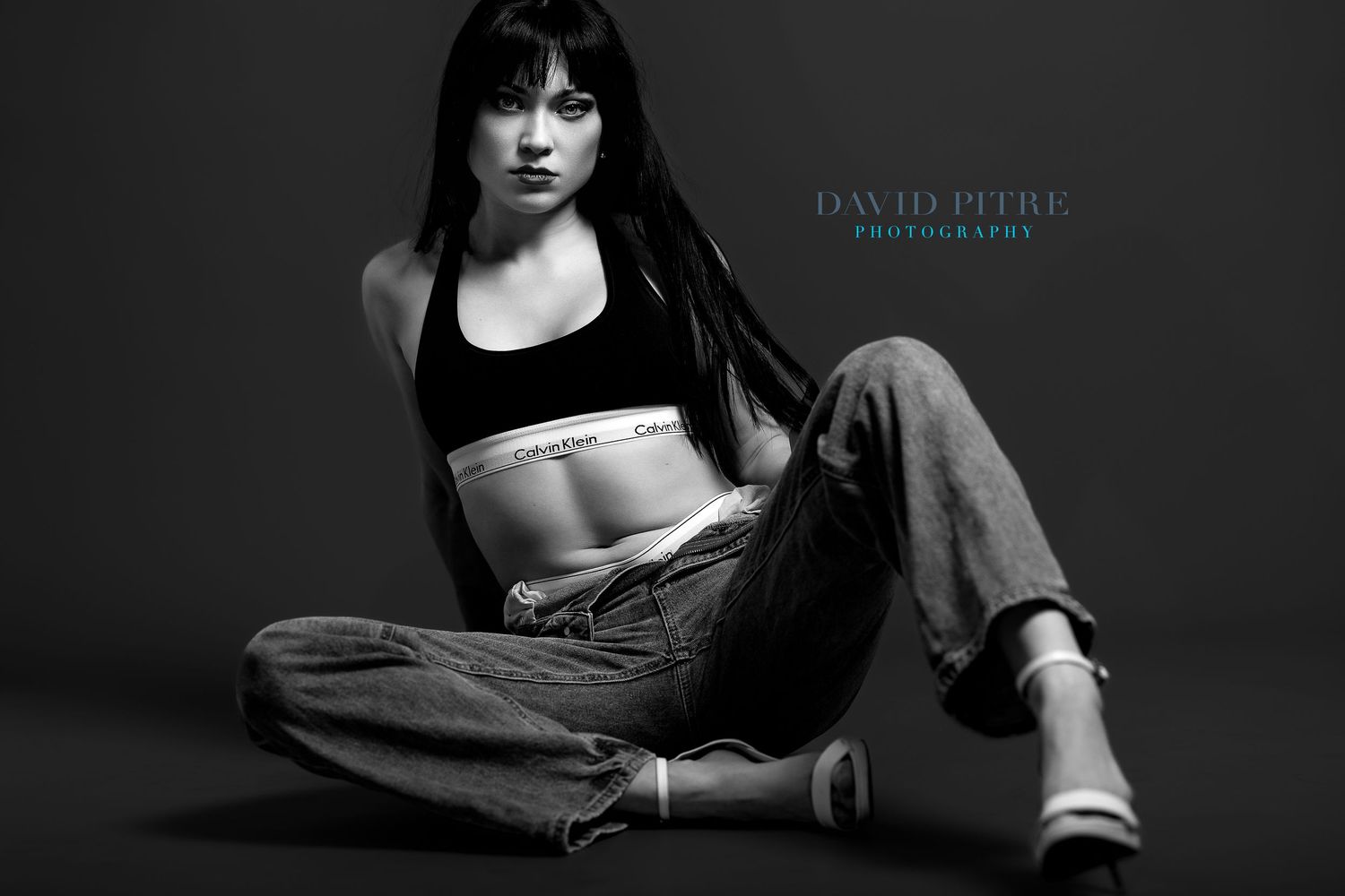 Black and white fashion photo showing athletic figure in crop top and baggy jeans sitting on dark floor.