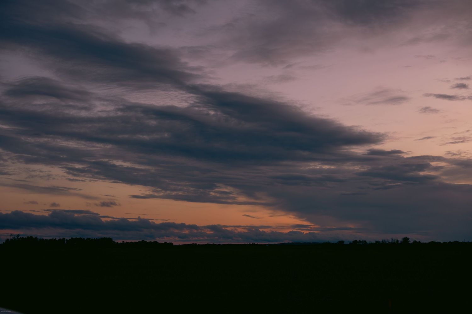 Dramatic dark clouds stretch across a moody sunset sky over a silhouetted rural landscape.