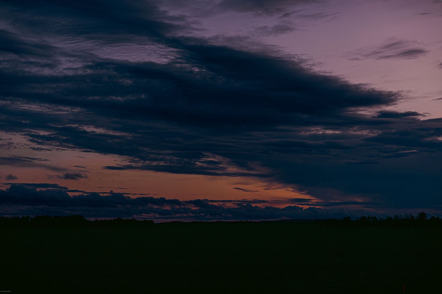 Panoramic view of a dark purple and orange sunset sky over a silhouetted treeline on the horizon.