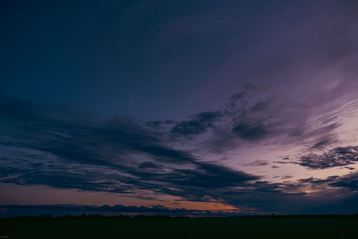 A dramatic purple and orange sunset sky with silhouetted clouds stretching across the horizon over a dark landscape.