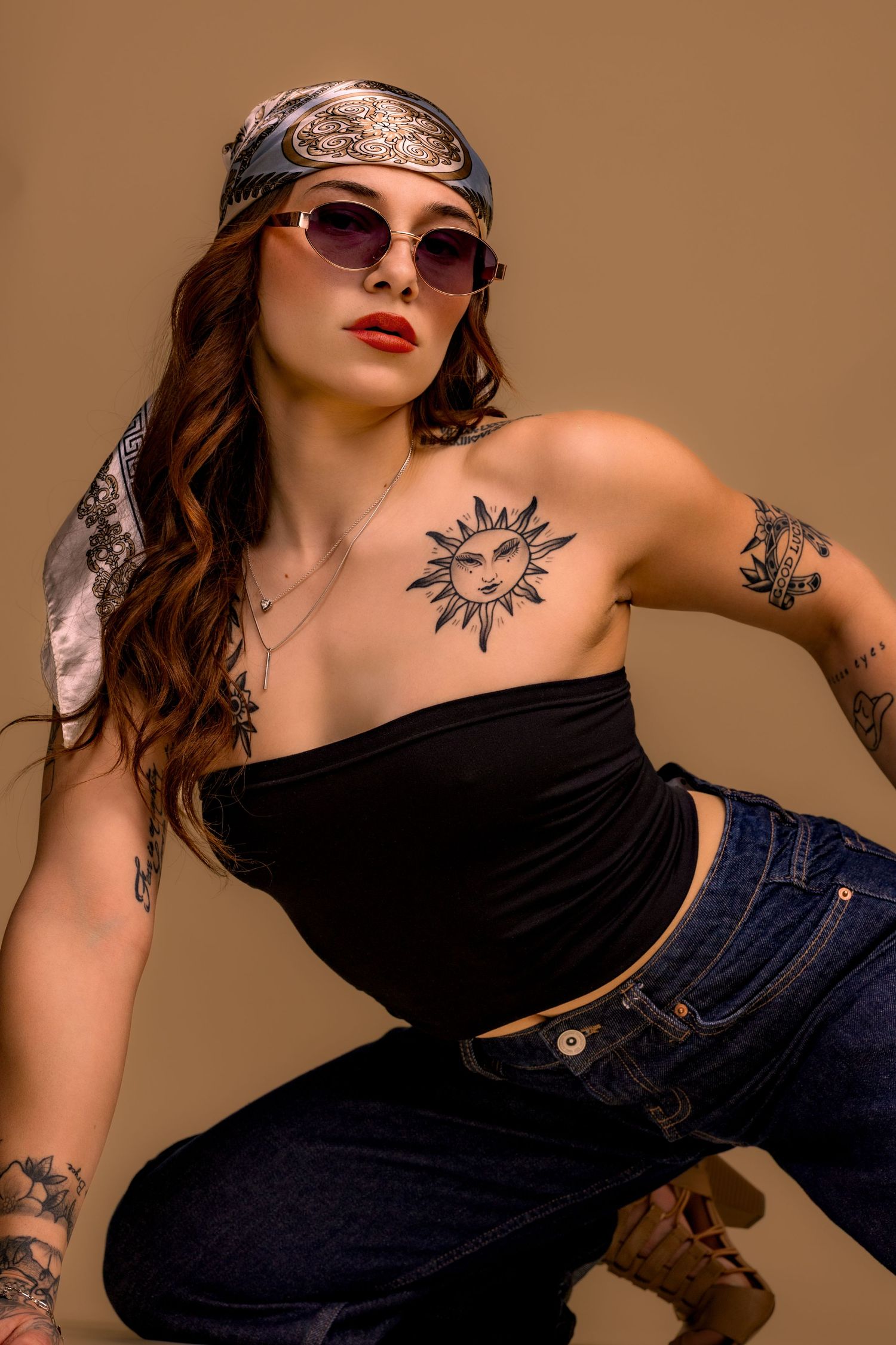A model in sunglasses and bandana poses in black crop top and jeans, showing artistic sun tattoo on shoulder.