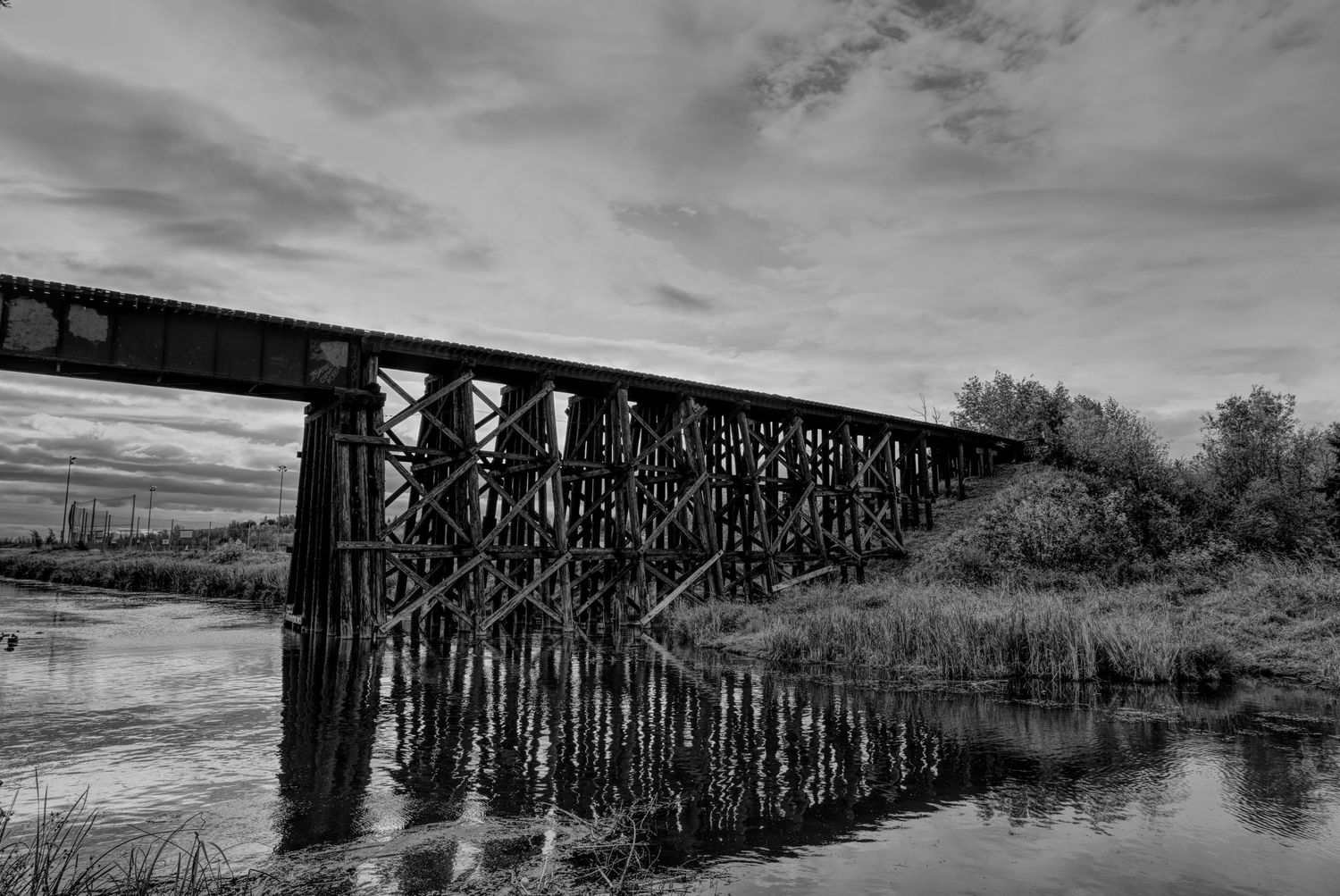 A dramatic black and white panorama of an old wooden railroad trestle bridge stretching across a calm river at sunset.