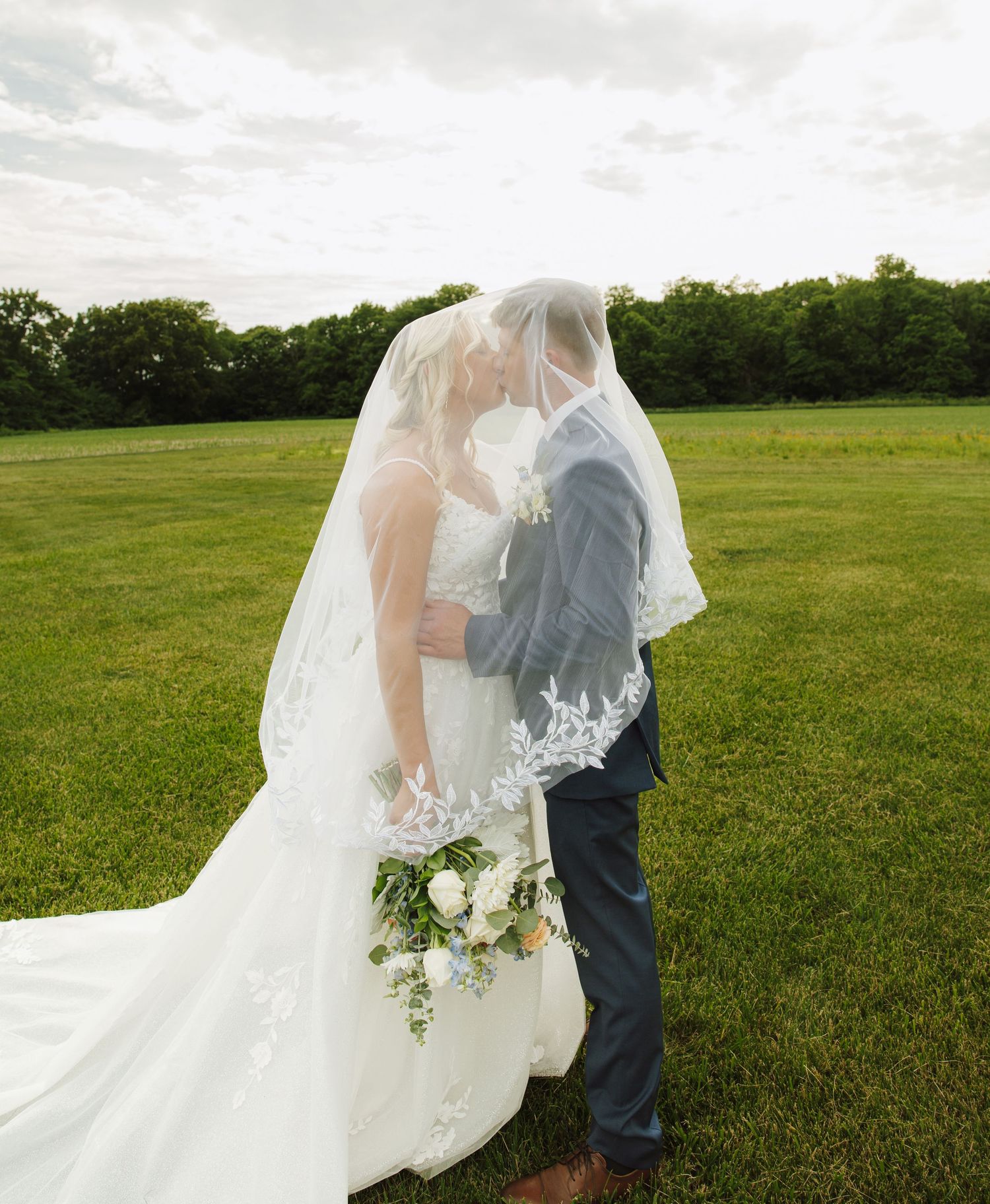Bride and groom kissing under lace wedding veil in green field on cloudy day.