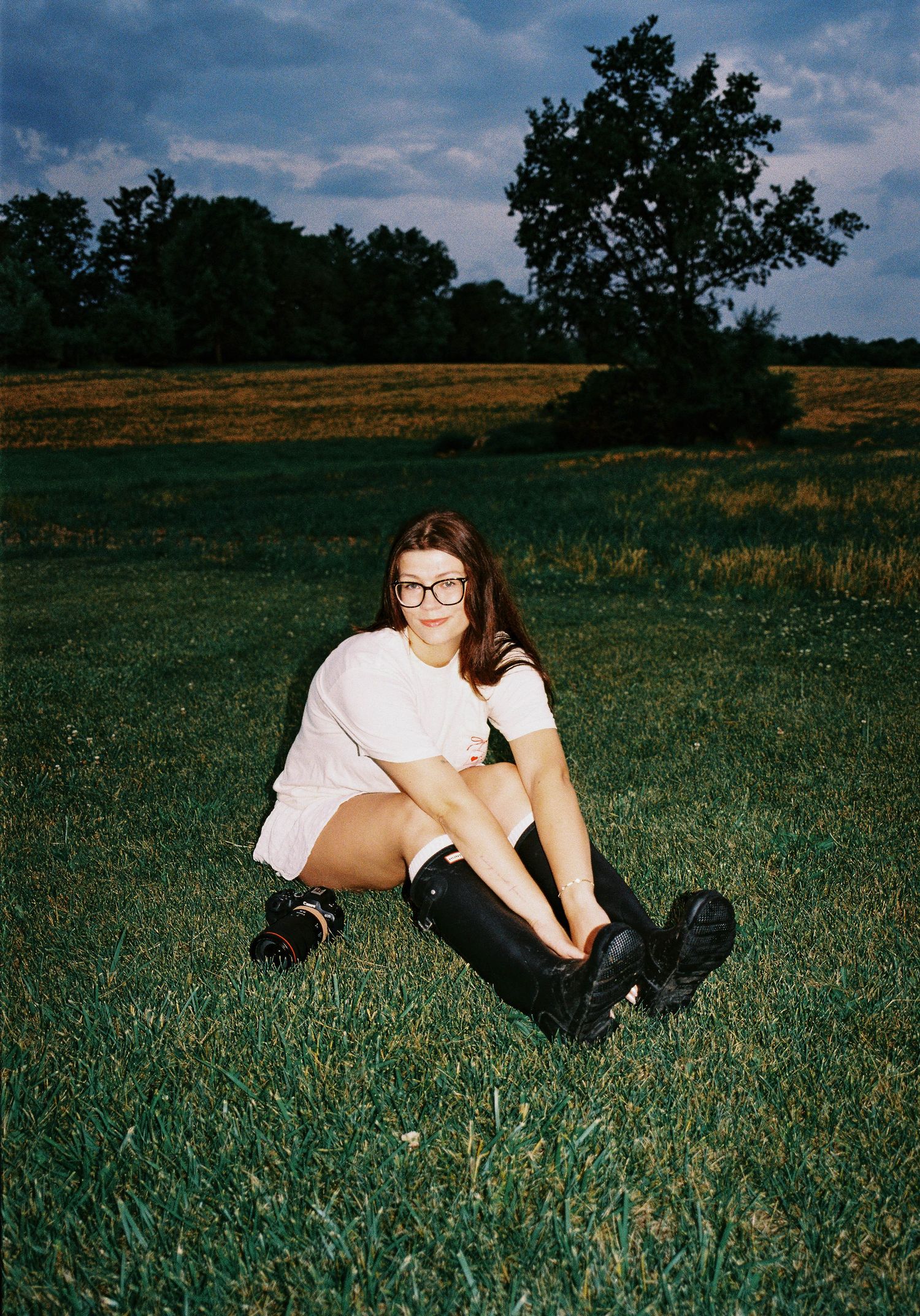Young woman in white outfit and black knee-high boots sitting in lush green grass field.