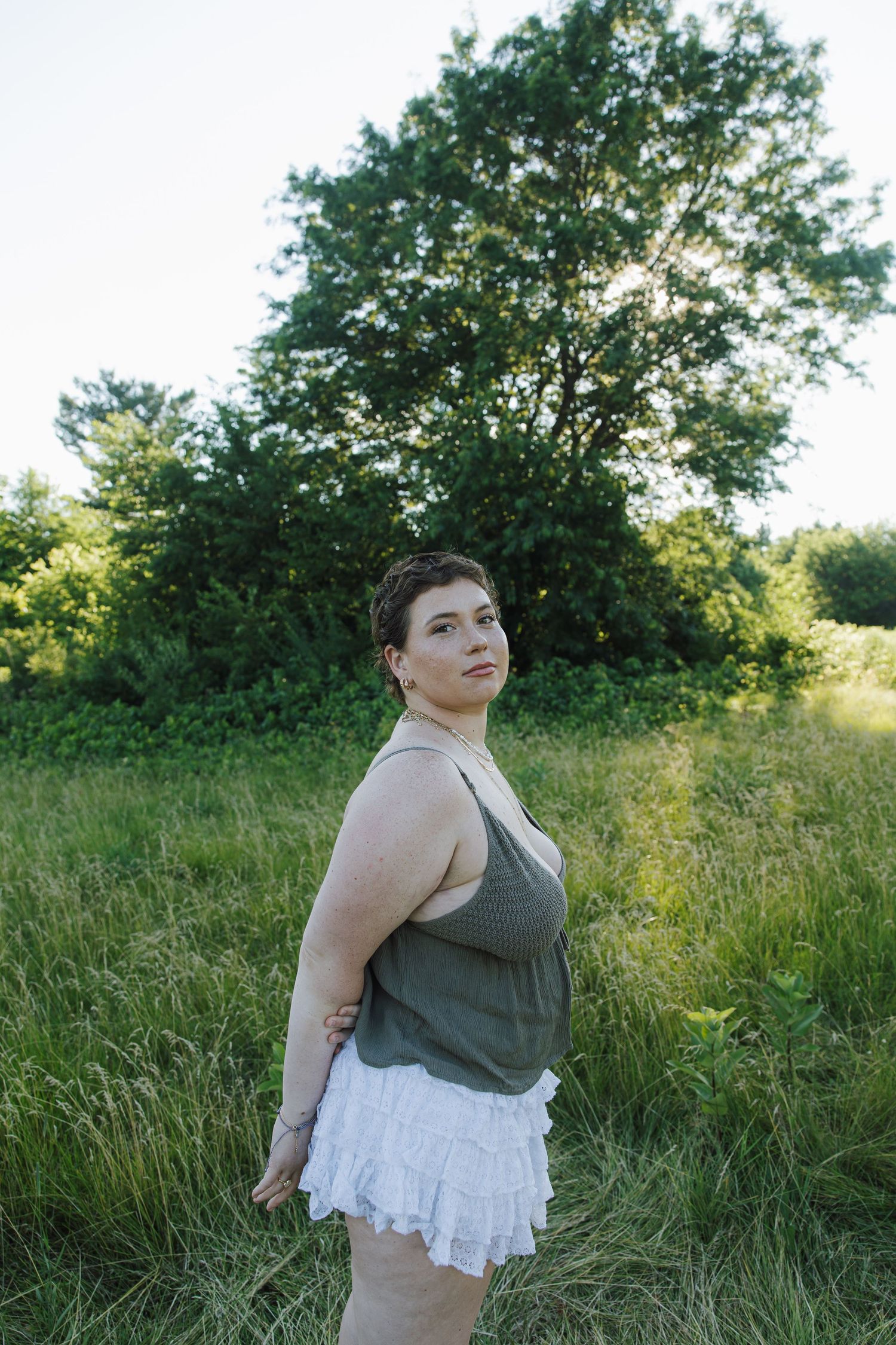 Woman in olive tank top and white skirt standing sideways in sunny green meadow.