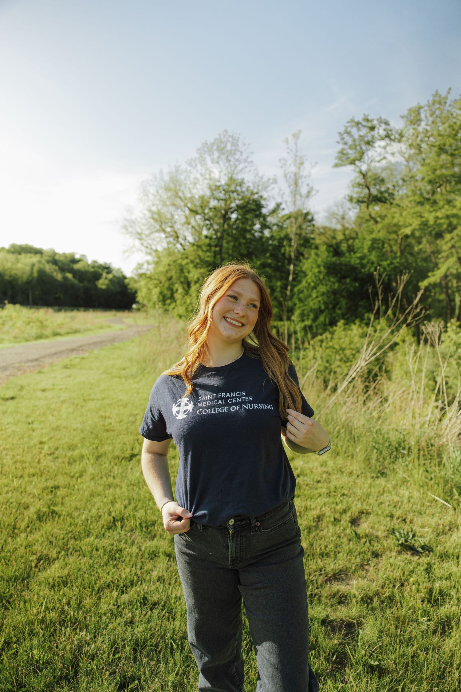 Smiling redhead woman wearing Saint Francis Medical Center College of Nursing t-shirt in green field.