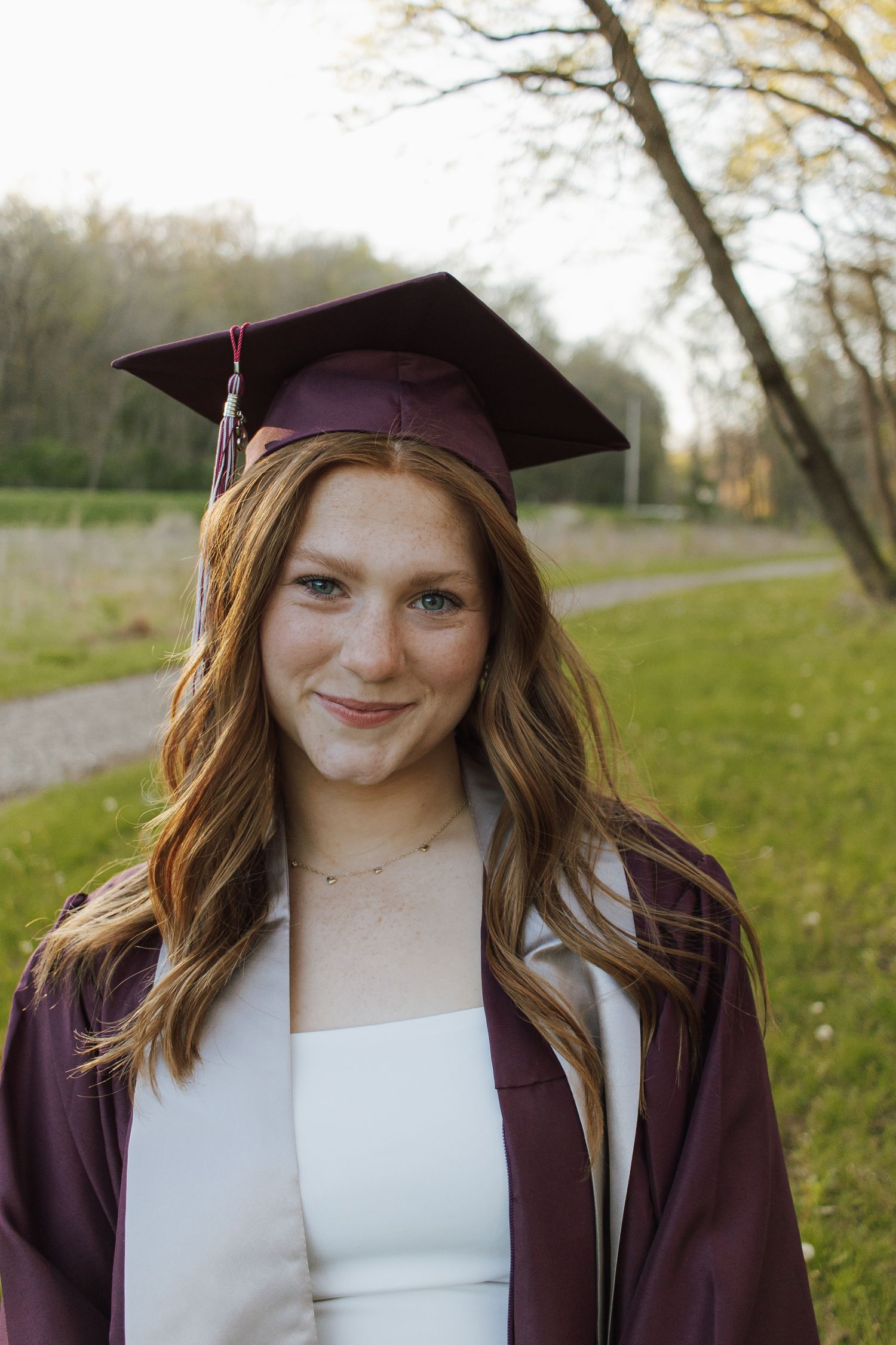 Smiling graduate in maroon cap and gown with white dress in outdoor park setting.