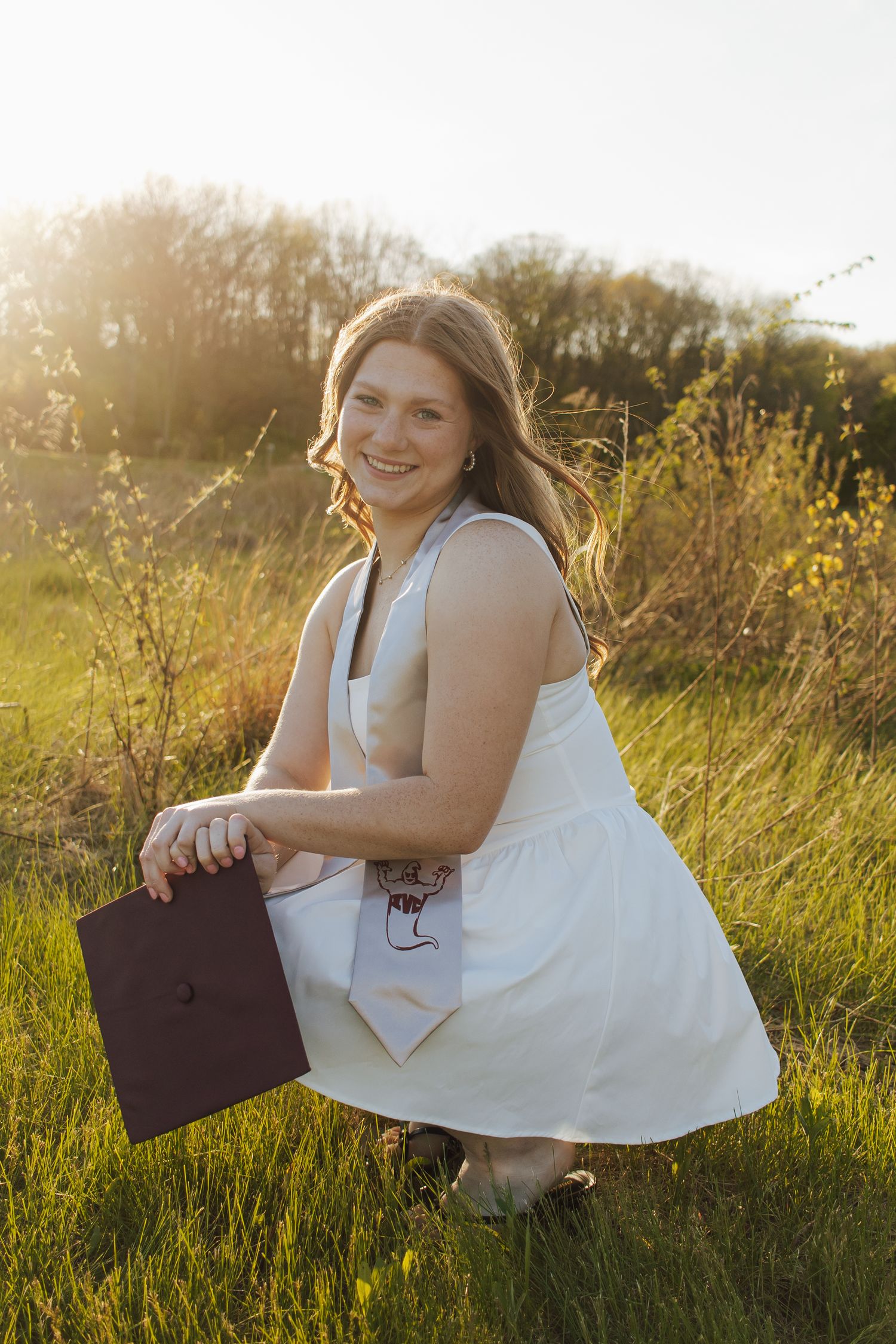 Young woman in white dress crouching in sunny field holding maroon graduation cap with stole.