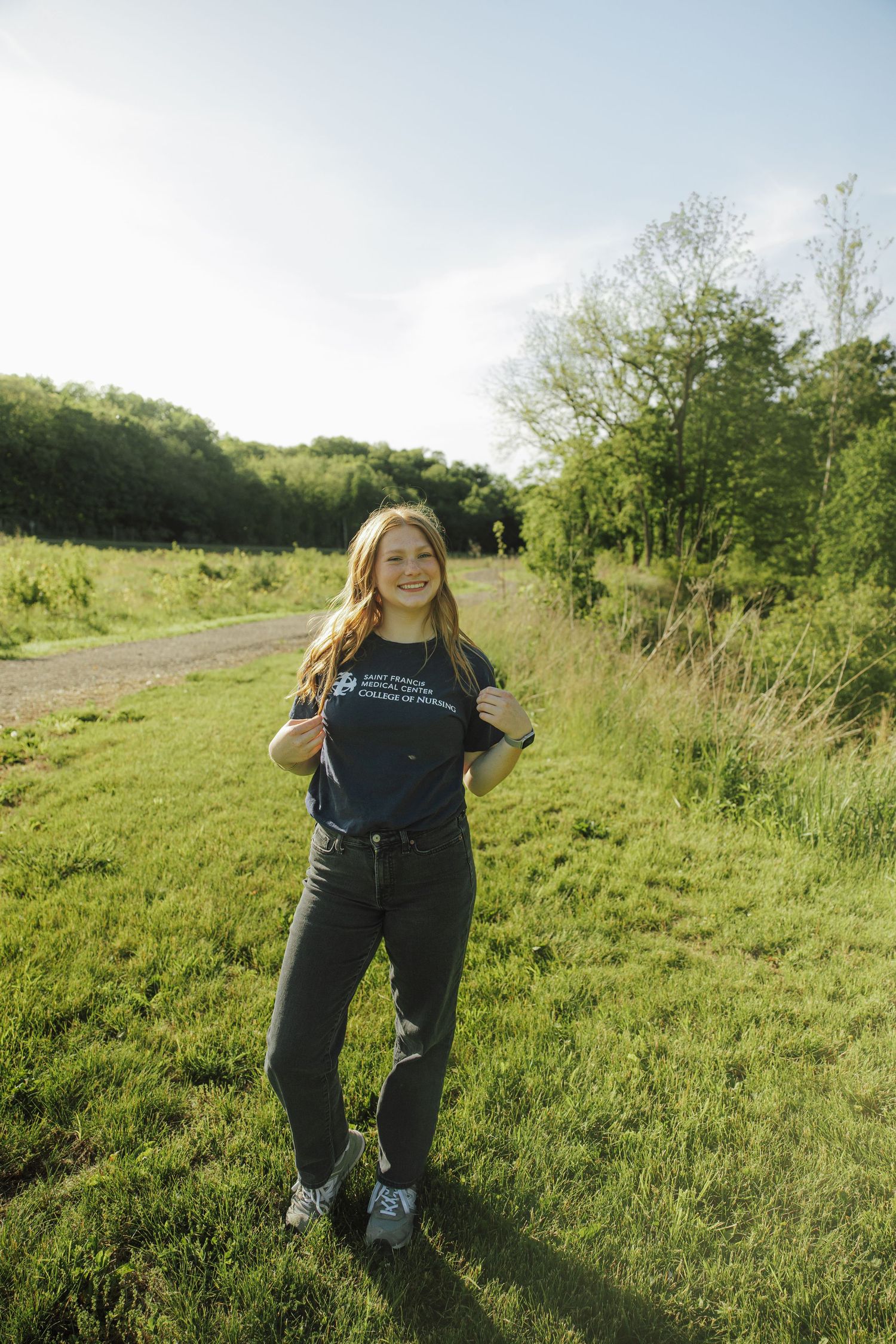 Smiling woman in nursing college t-shirt standing on grassy path with trees in background.