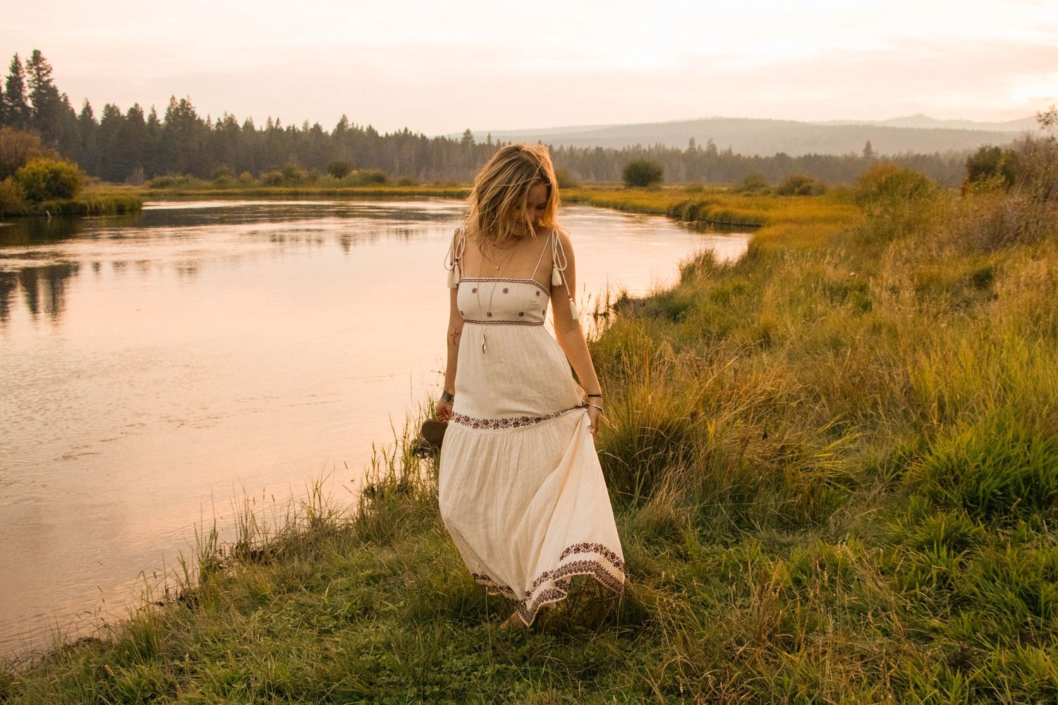 a young woman stands beside a river looking down at the grass at a park in Bend, Oregon