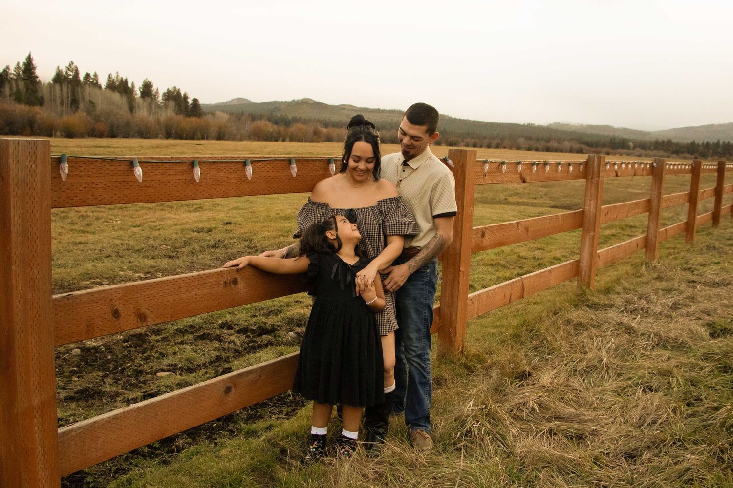 a mom and dad lean against a rustic wood fence in Sisters, Oregon, with a horse pasture beside them