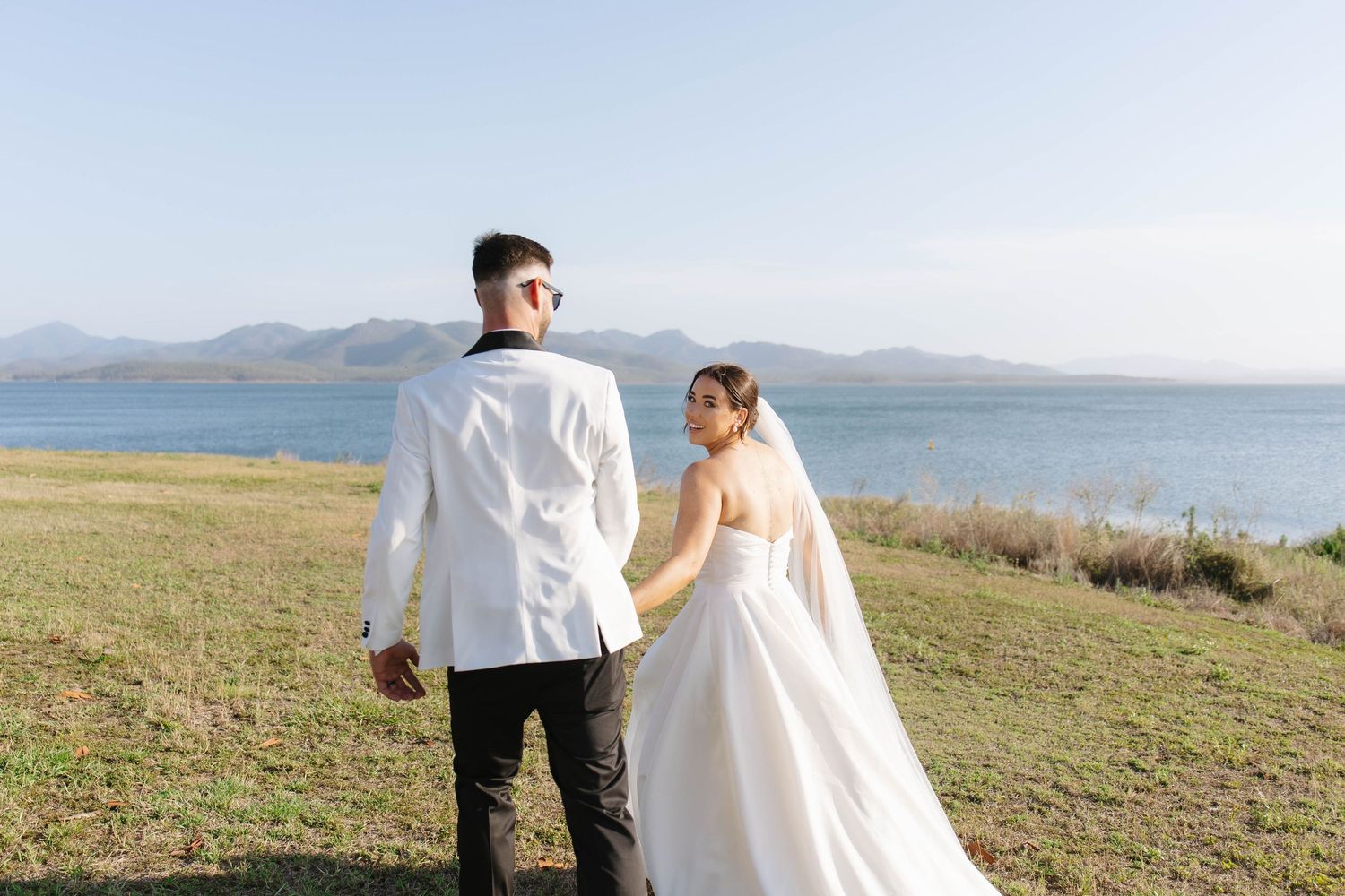 A couple in wedding attire stands on a grassy hillside overlooking a scenic lake and mountains.