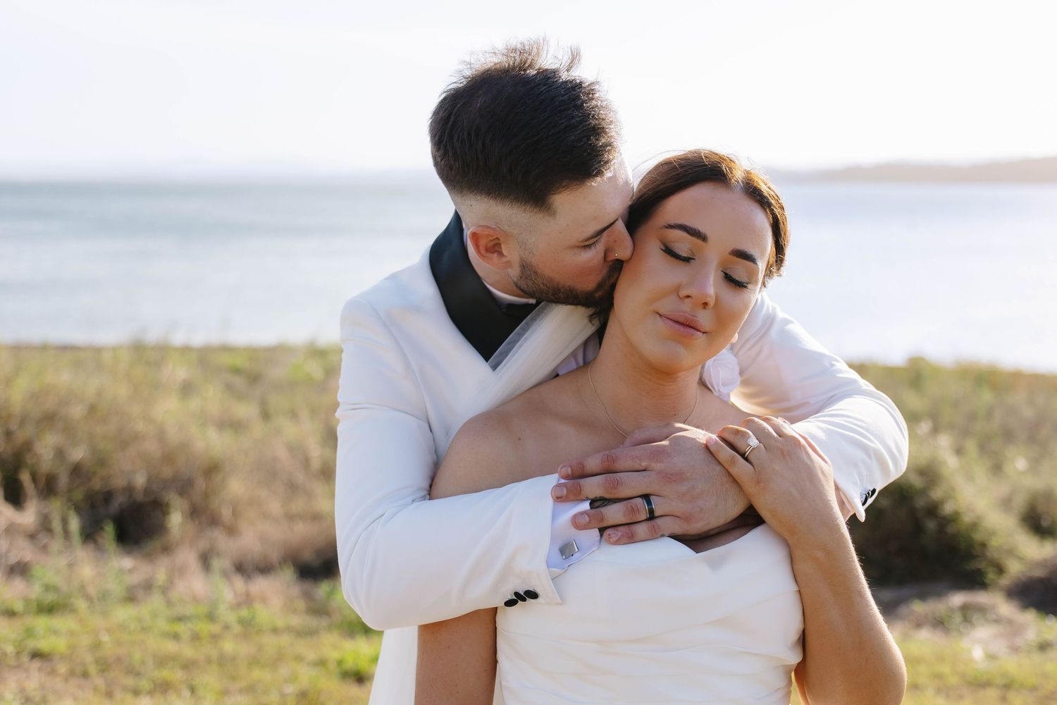 A romantic wedding couple embraces on a hillside with a serene lake view in the background.