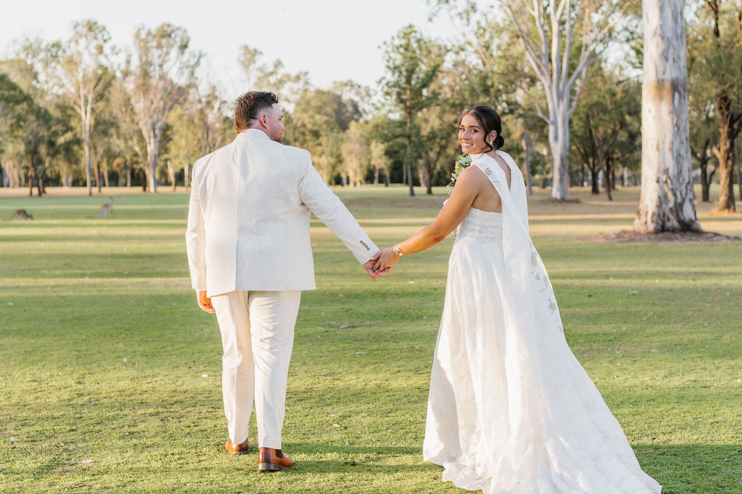 Newlyweds in white walk hand-in-hand across a scenic park lawn.