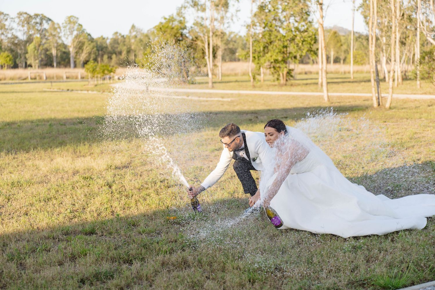 Playful newlyweds splash through sprinklers on a sunny field during their wedding celebration.
