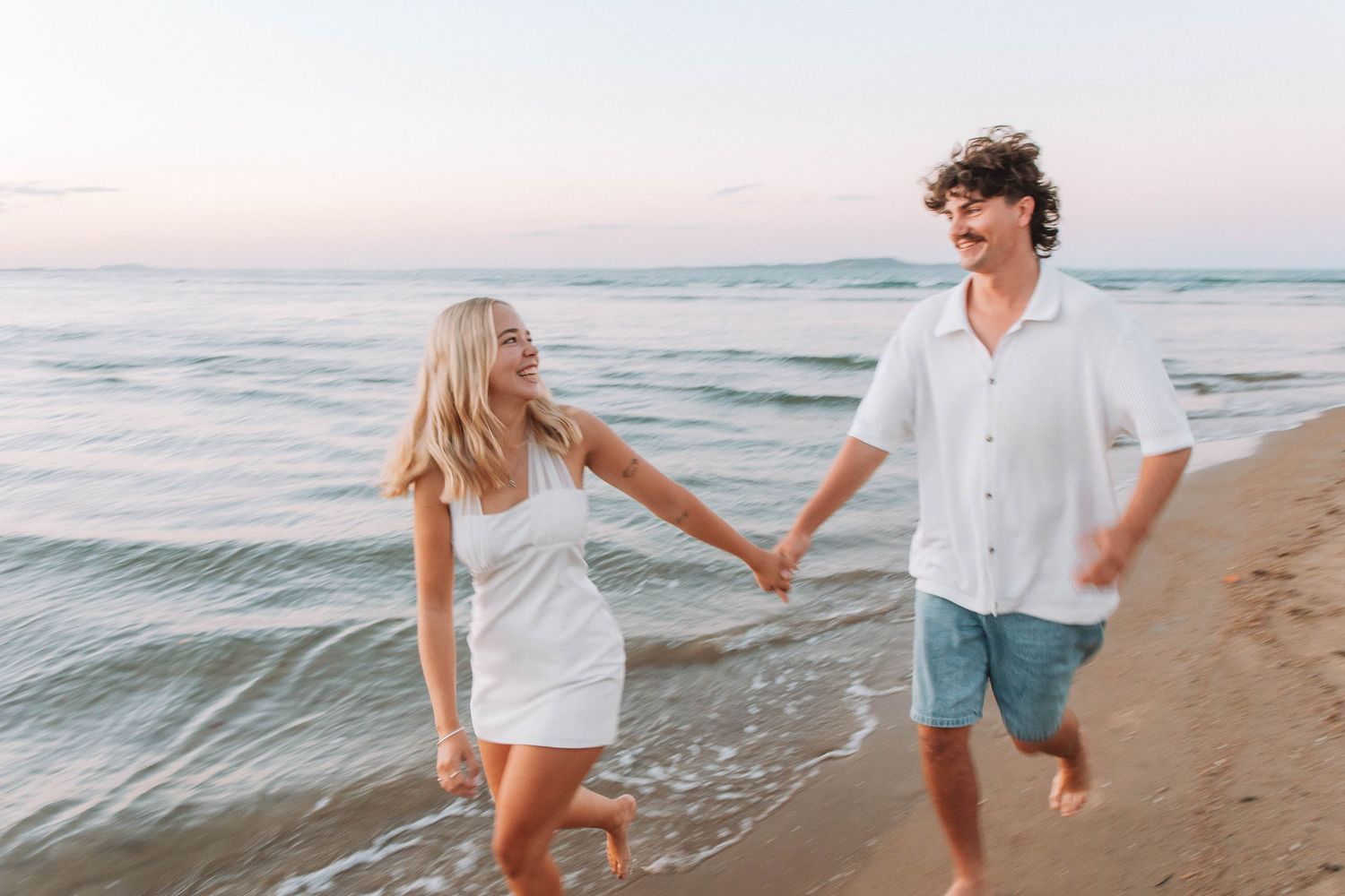 A happy couple holds hands while running along a sandy beach at sunset, wearing casual white summer outfits.