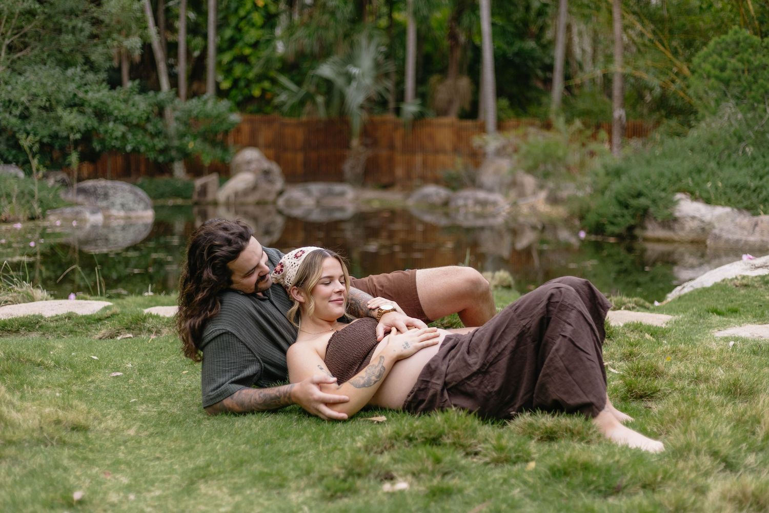 A couple embraces while lying in a lush green garden setting on a sunny day.