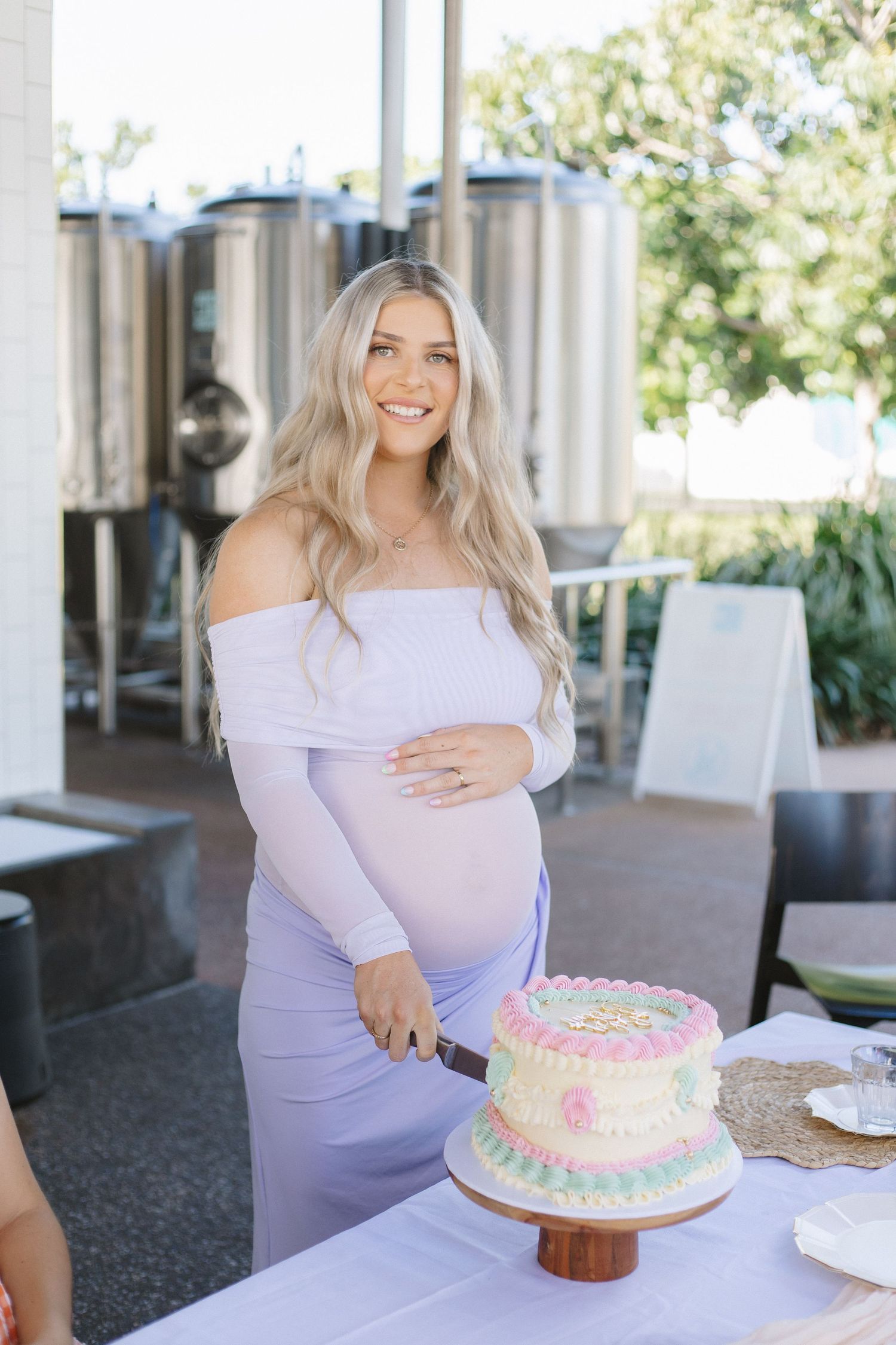 A person in a lavender dress poses with a decorated birthday cake at an outdoor celebration.