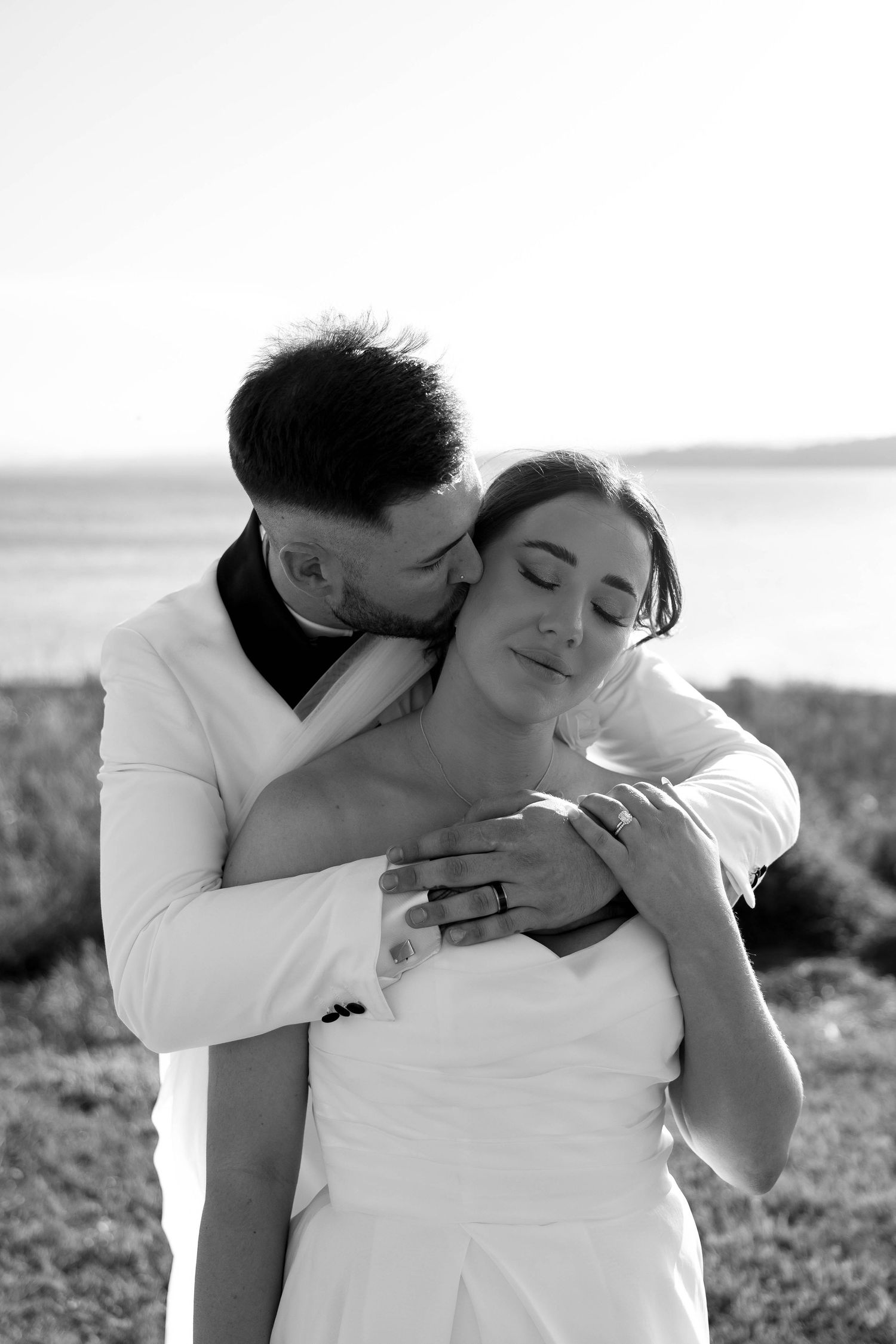 A black and white portrait captures an intimate moment between newlyweds against a waterfront setting.